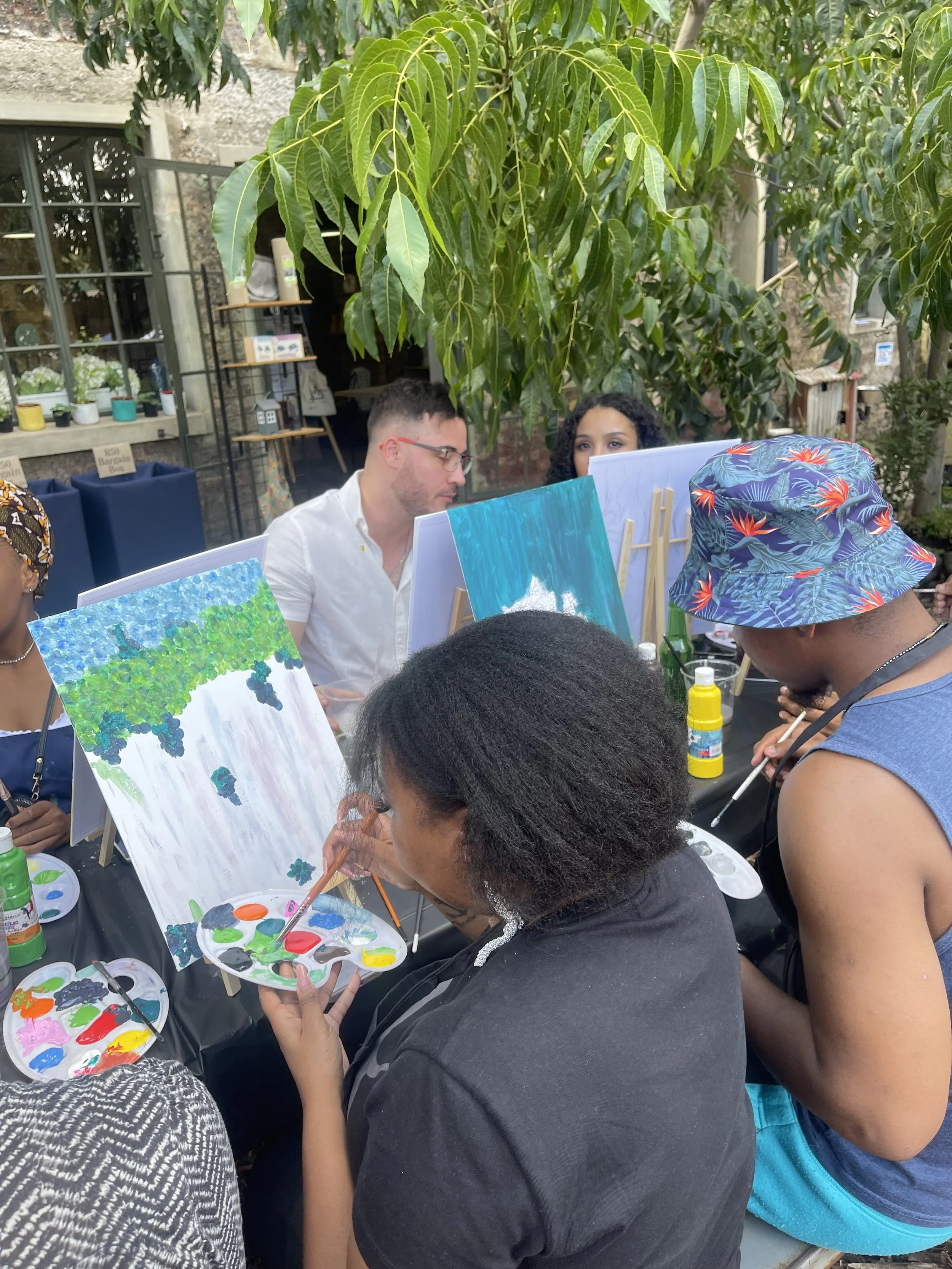A group of people painting small canvases with outdoor and nature themes at a craft table during a social event.