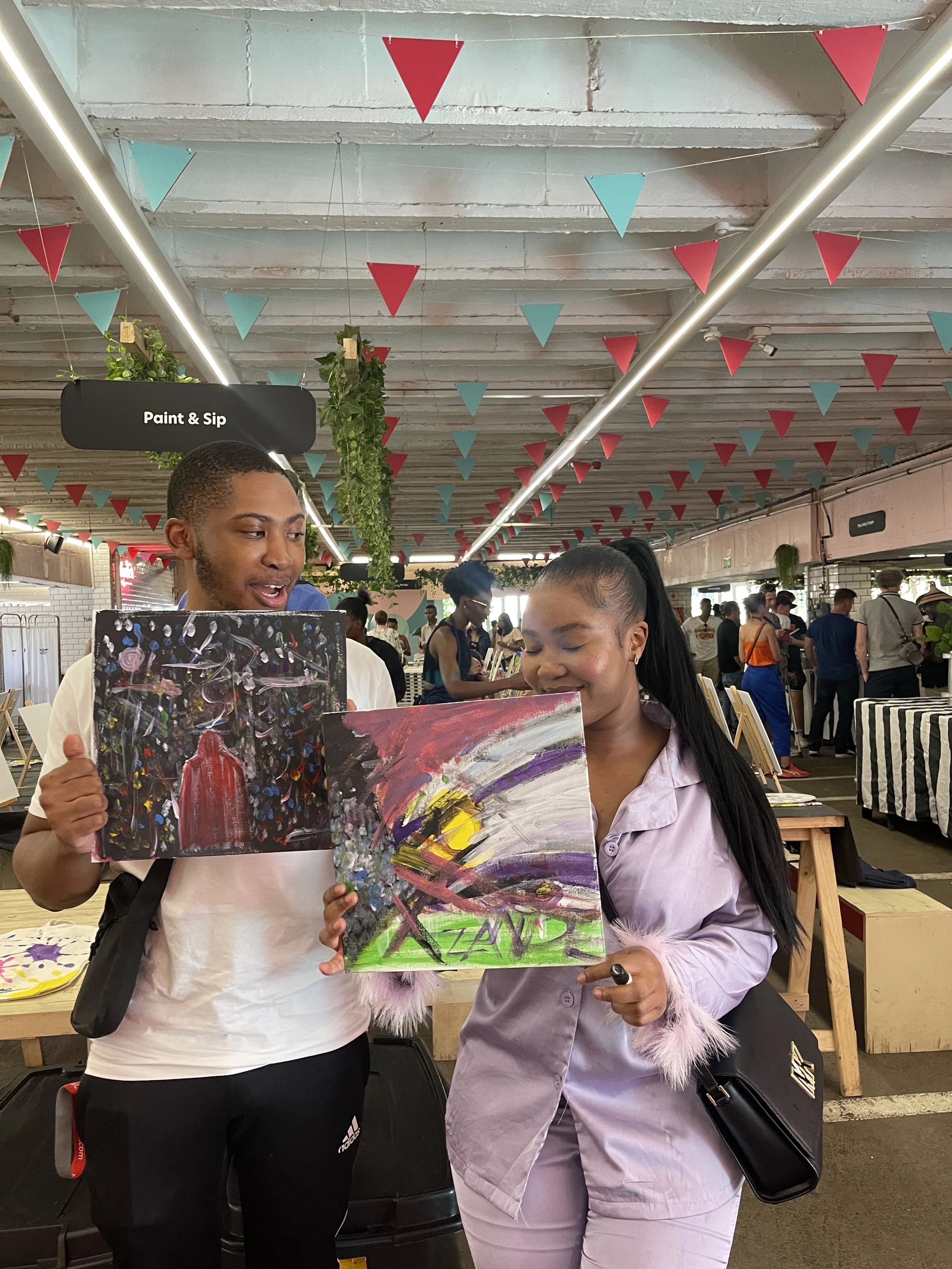 Two people, a man and a woman, are holding colorful abstract paintings in an indoor setting with a decorated ceiling featuring red and teal triangular banners and green hanging plants. There are many people in the background.
