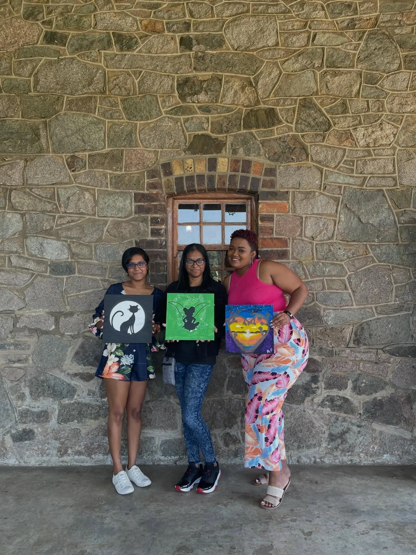 Three women standing in front of a stone wall, each holding a painting of a cat, fairy, and sunset heart.
