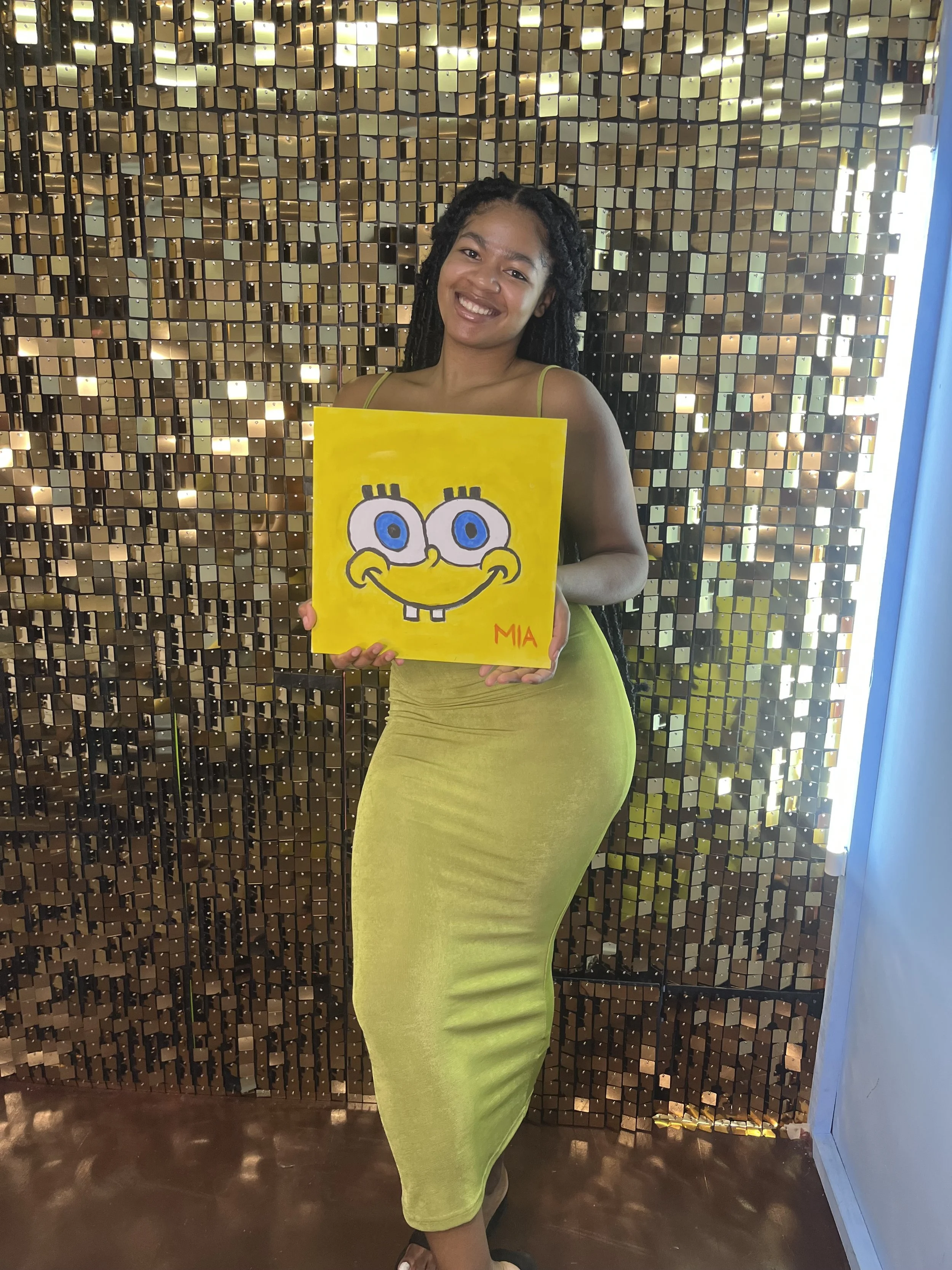 A woman with dark curly hair, smiling, holding a yellow drawing of SpongeBob SquarePants in front of her, standing in front of a shiny, gold-colored, textured wall.
