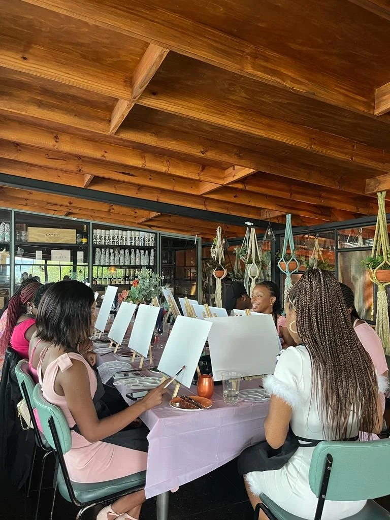 Women participate in a painting class at a restaurant with a wooden ceiling, painting on canvas strips, seated around a table with pink tablecloths.