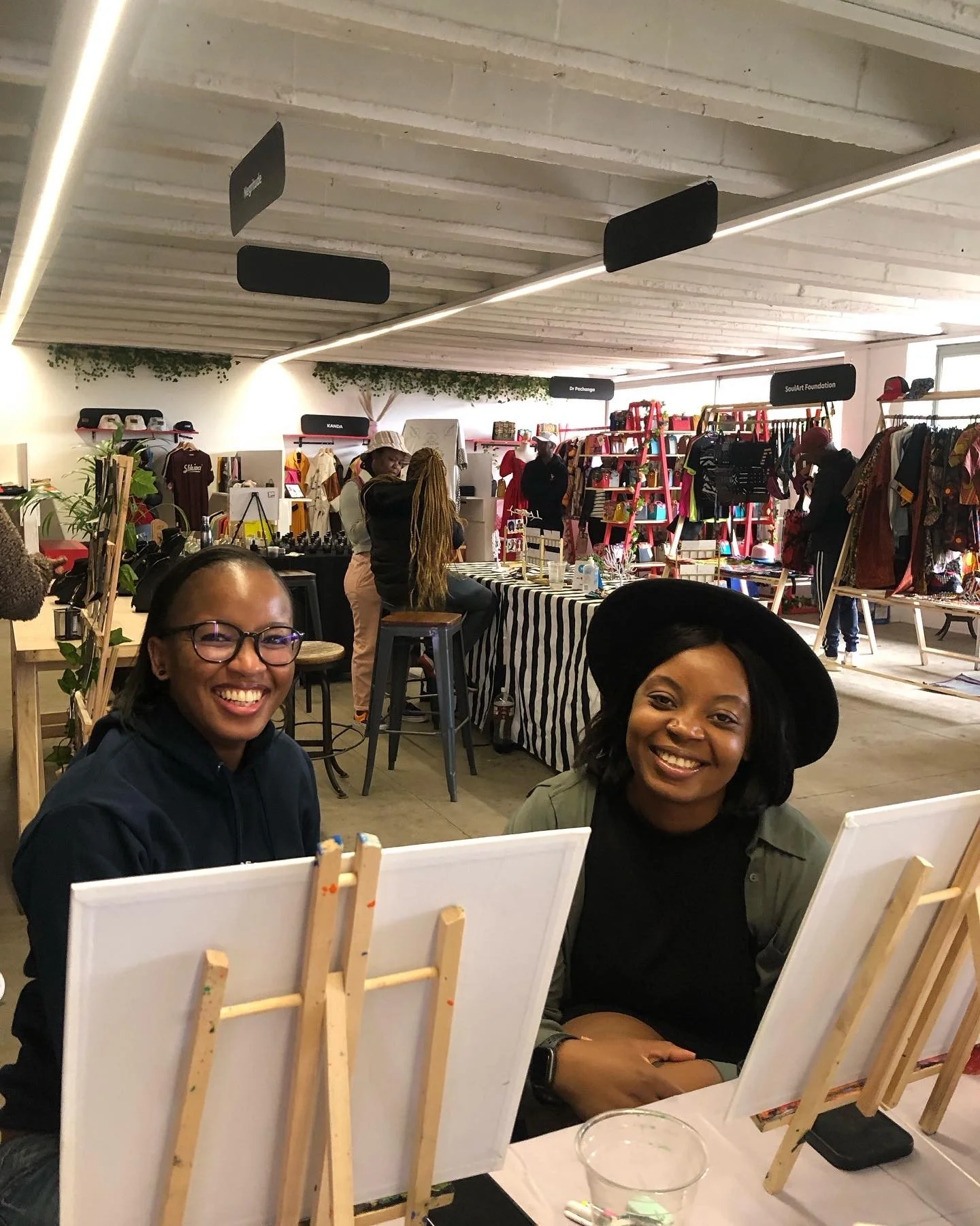 Two women smiling at an art event, sitting in front of canvases and easels with paint supplies. The setting is a well-lit indoor space with display racks and other attendees in the background.