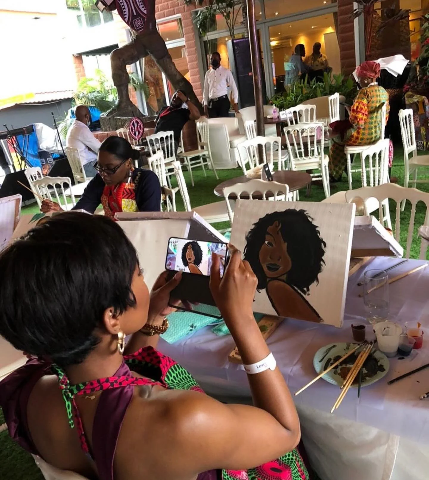 A woman is painting a portrait of a woman with curly hair at an art event, while another woman is taking a photo of the painting with her phone. Several people are sitting and standing outside under a tent, some looking at their phones and others obs