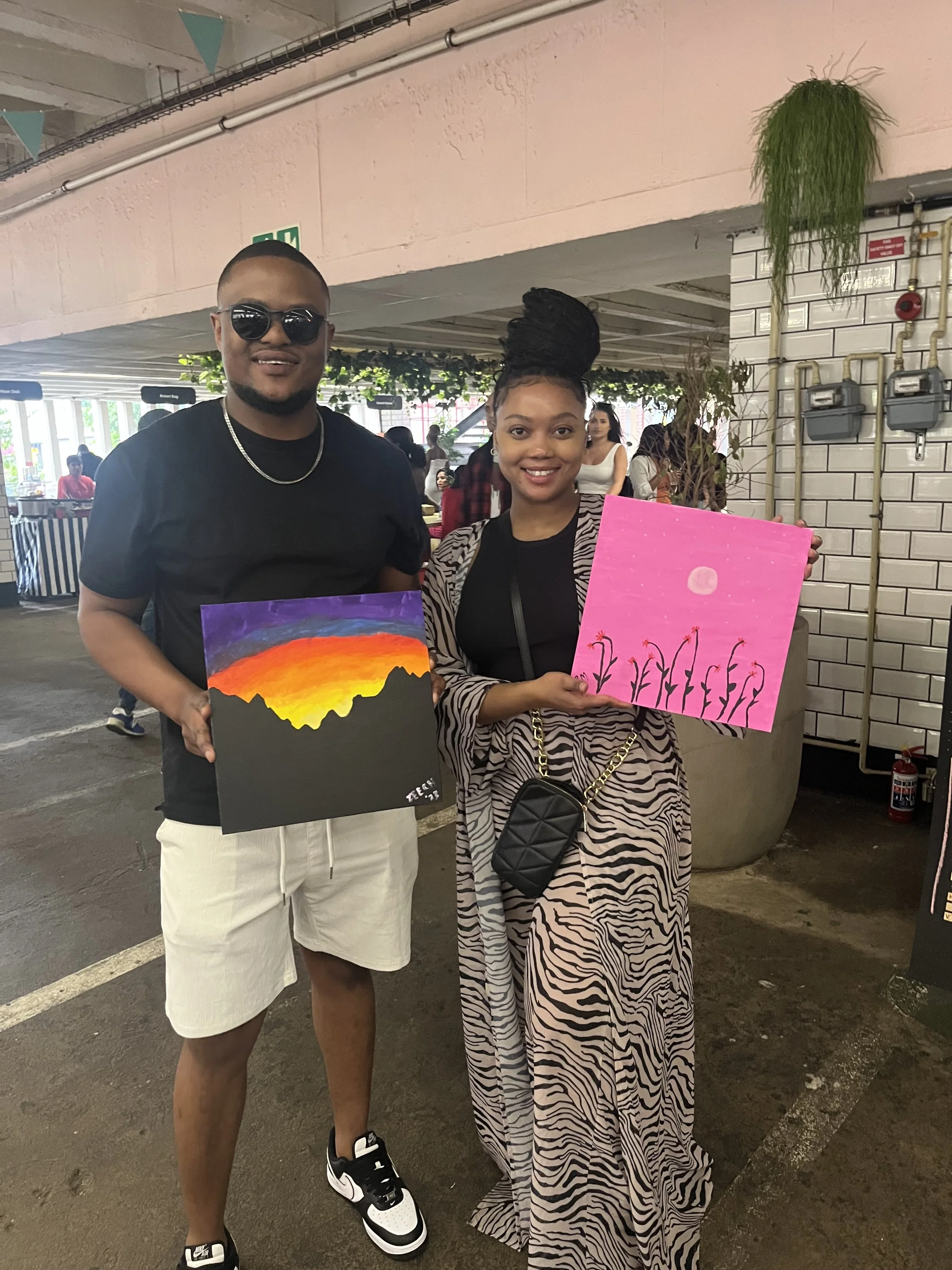 A man wearing sunglasses, a black t-shirt, and white shorts stands next to a woman with a high braided hairstyle, a zebra print dress, and a black crossbody purse. They are holding paintings with sunset and moon themes, in an indoor setting with othe