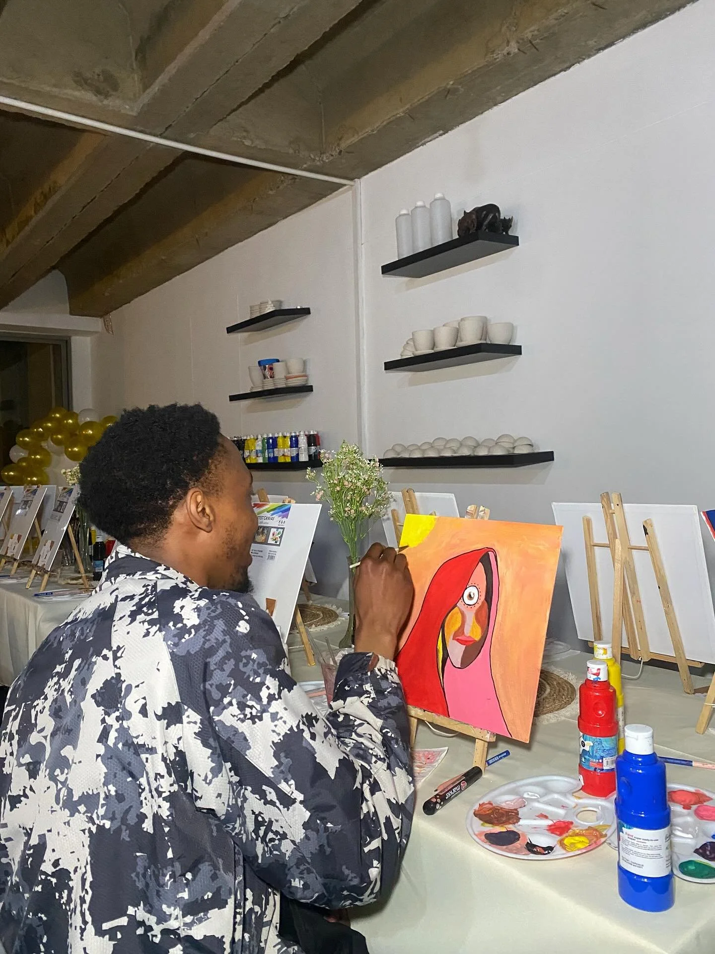 An artist painting a colorful portrait of a woman with long red hair on a canvas. The artist is wearing a black and white patterned jacket and is sitting at a table with art supplies, including paints and brushes. Behind them, there are shelves with cups, plates, and decorative items, as well as balloons and flowers in the background.
