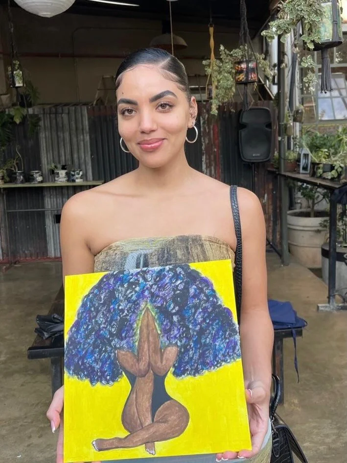 A young woman with dark hair and hoop earrings holding a colorful painting of a nude woman with an afro hairstyle, sitting with her knees pulled up and hands covering her face, against a yellow background, in an indoor art space with plants and indus