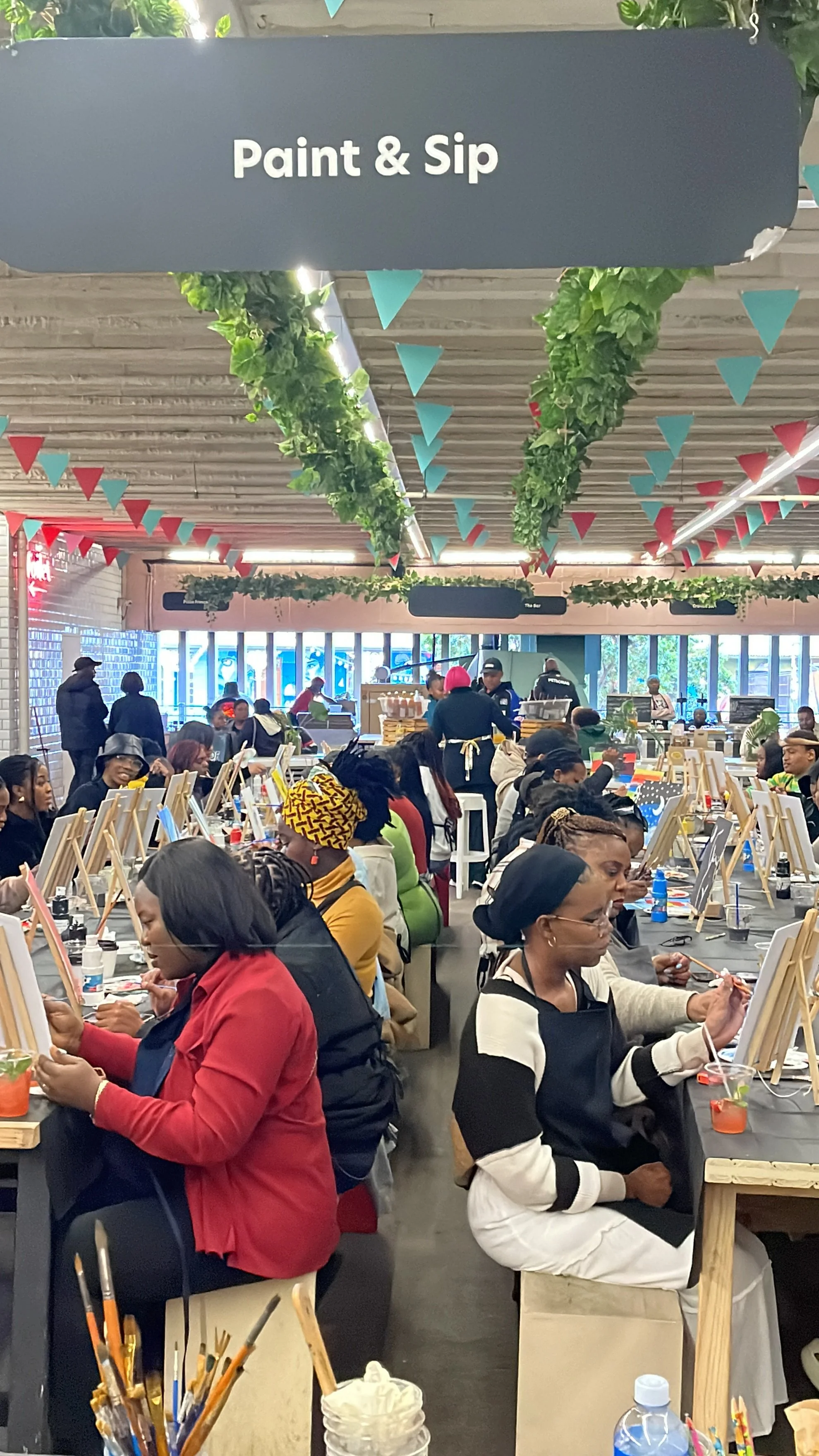 Indoor painting event with many people seated at tables, each with an easel and art supplies, under a sign that says 'Paint & Sip' with colorful decorations hanging from the ceiling.