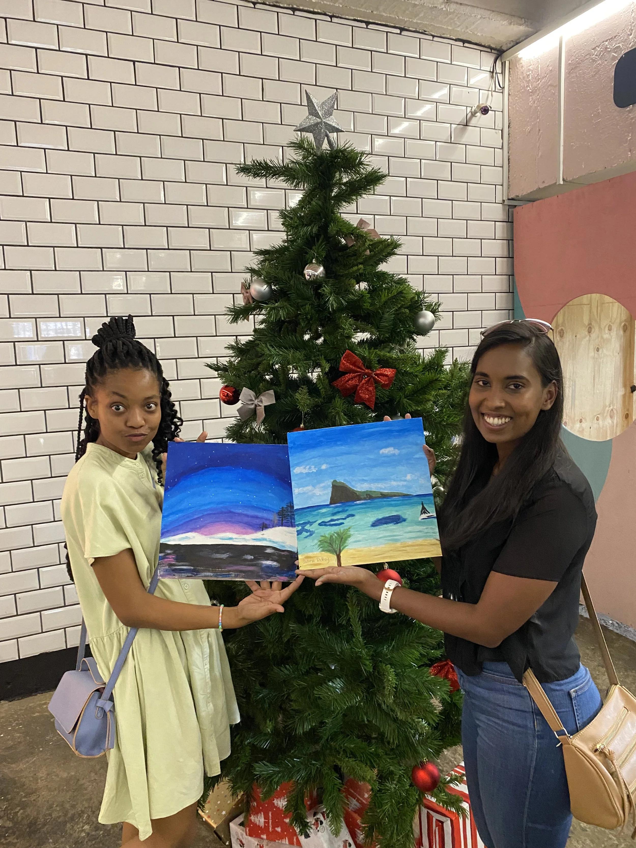 Two women holding a painting of a beach scene with a mountain in the background, standing on either side of a decorated Christmas tree with wrapped gifts underneath, against a white-tiled wall.