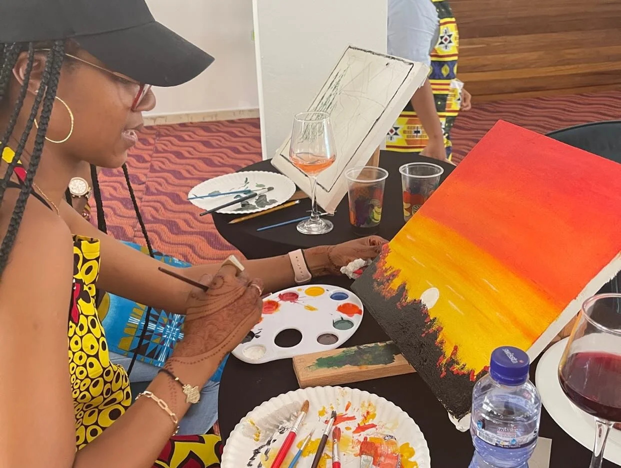 A woman painting a sunset on a canvas at a table with wine and painting supplies during an art event.