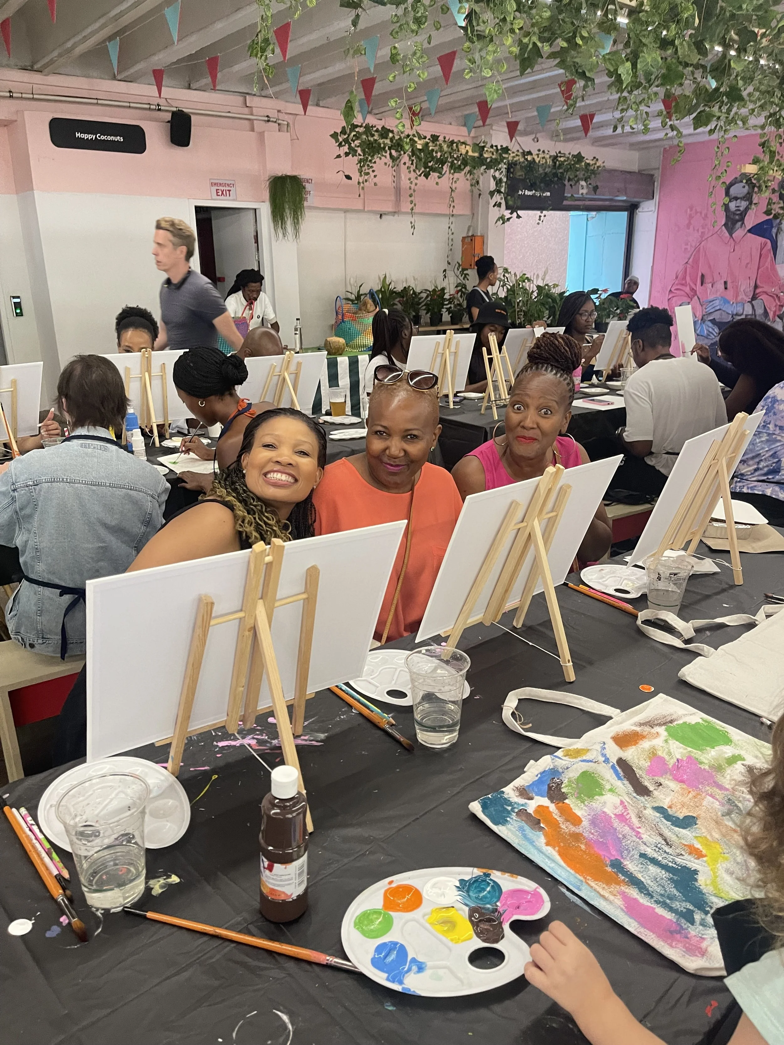 Group of people participating in a painting activity, with easels, paints, and brushes on a long table covered with black plastic. Some individuals are focusing on their artwork, while three women smile at the camera in the foreground. The room has pink and white walls, hanging greenery, and colorful triangular pennant banners overhead.