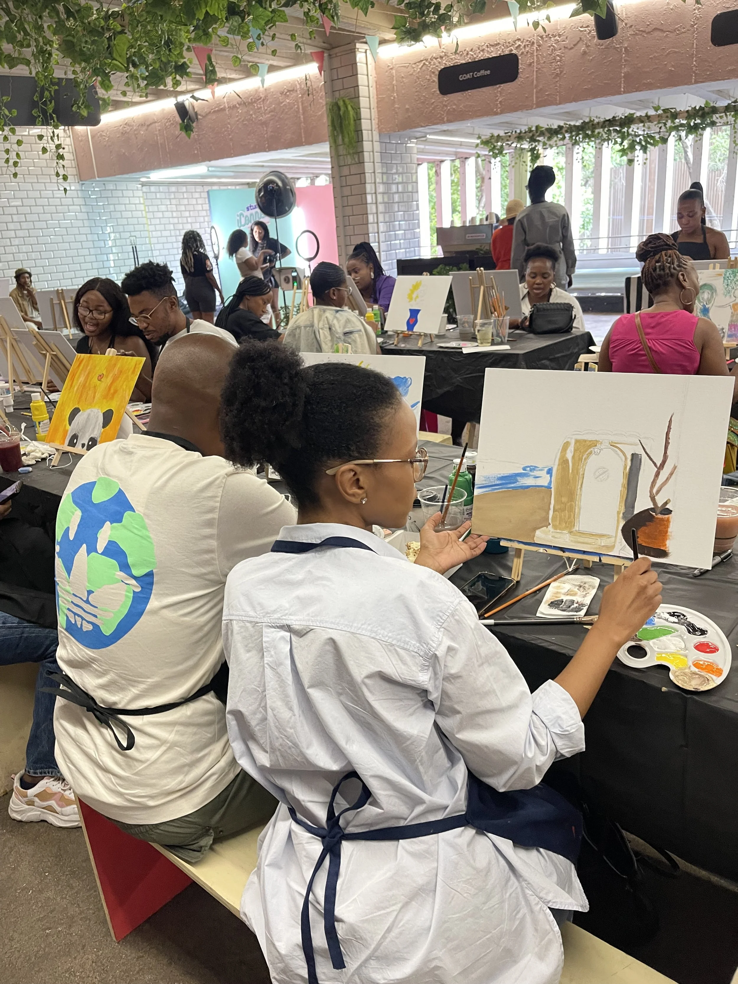 People participating in a group painting event indoors, with canvases, paints, and brushes on tables, some wearing aprons, and paintings of animals and Earth on display.