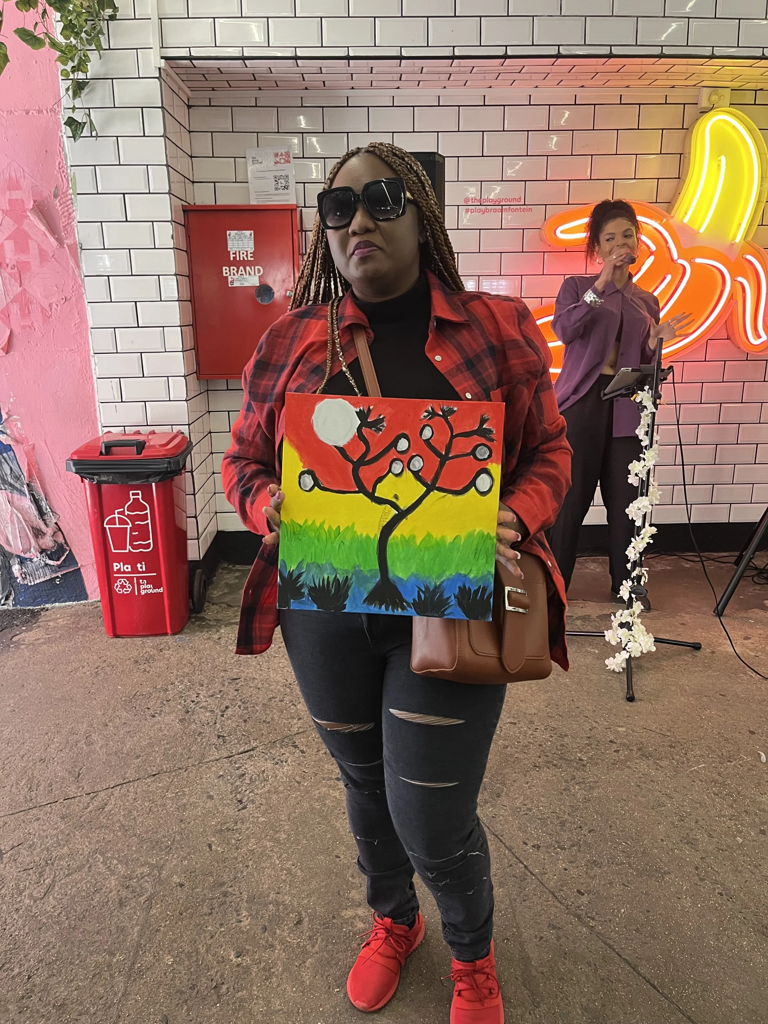 A woman with braided hair wearing black sunglasses, a red and black plaid shirt, and ripped jeans is holding a colorful painting of a tree with a white moon in the sky. She is standing in front of a white brick wall with a neon banana sign and a woma