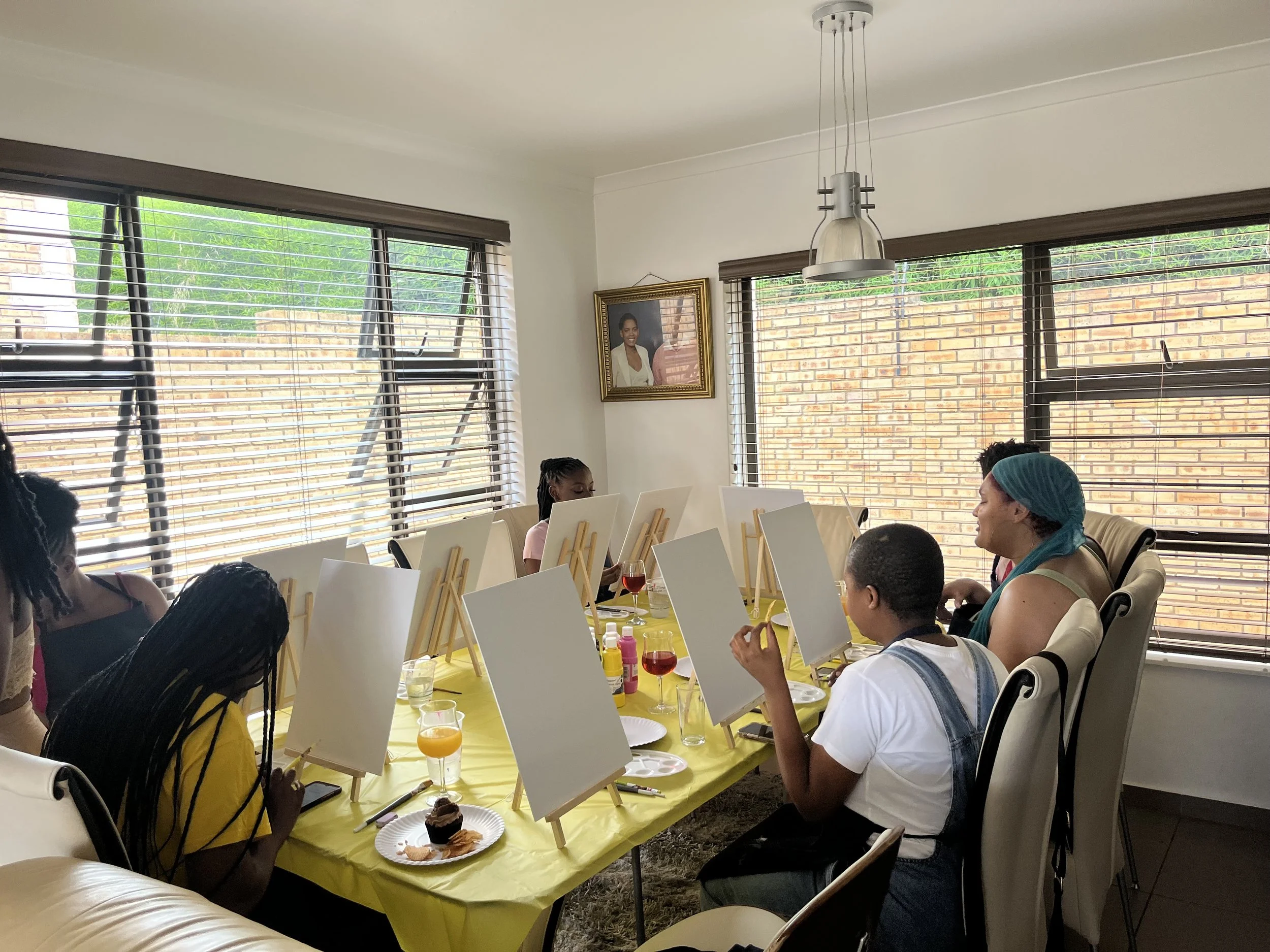 People sitting at a table with blank canvases on easels, paint, and drinks, participating in an art class or workshop in a bright room with large windows and a photo on the wall.