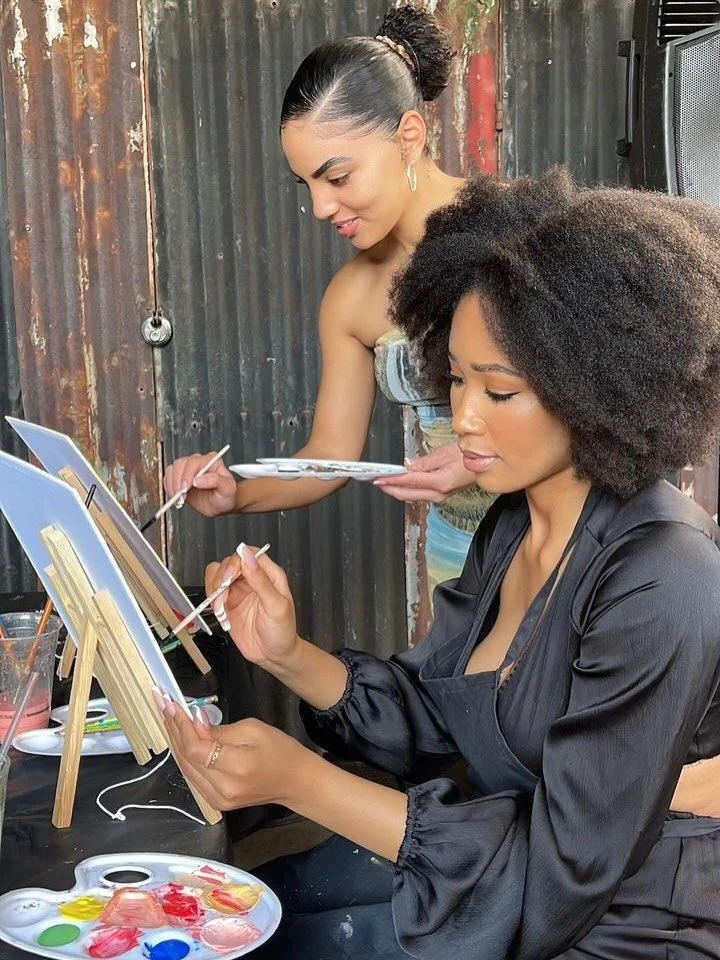 Two women painting on canvases, one sitting and one standing, outdoors with a rustic metal wall background.
