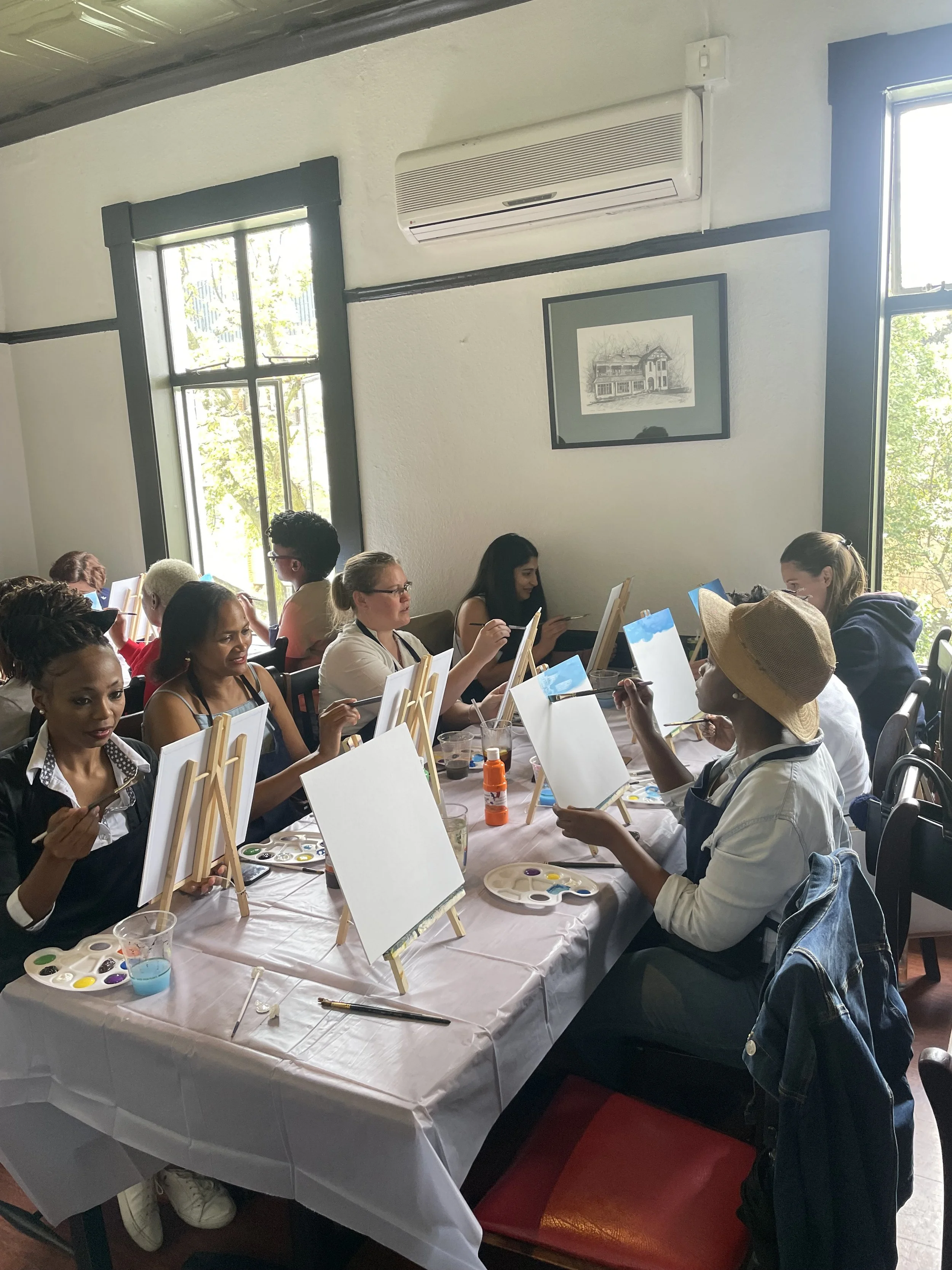 People participating in a group painting activity at a long table covered with a white tablecloth, with canvases, paint palettes, and brushes, in a room with large windows and framed artwork on the wall.