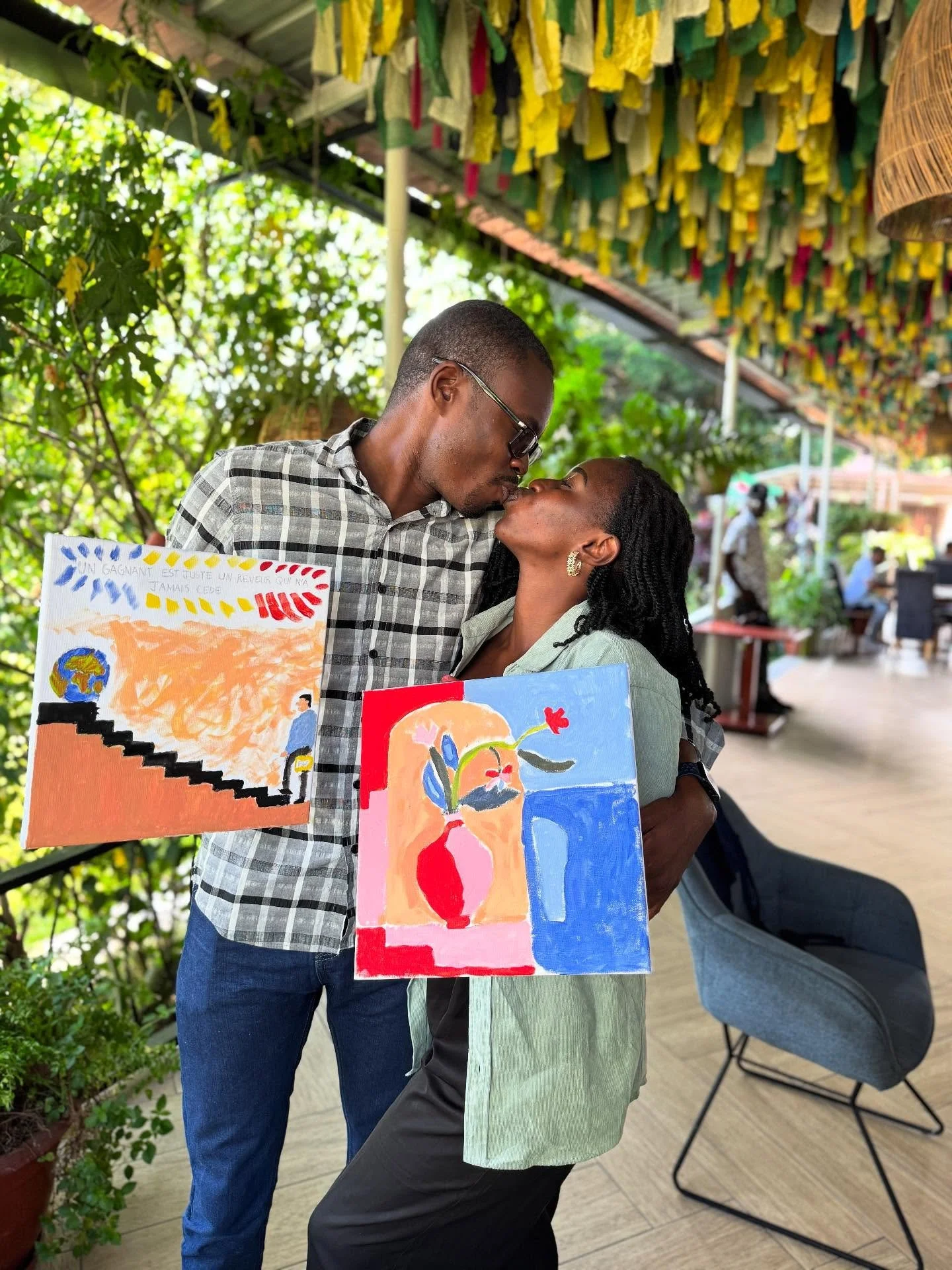 Two people sharing a kiss at an outdoor art event, holding paintings with colorful abstract designs and a drawing of the world. The background features hanging yellow and green fabric and lush greenery.