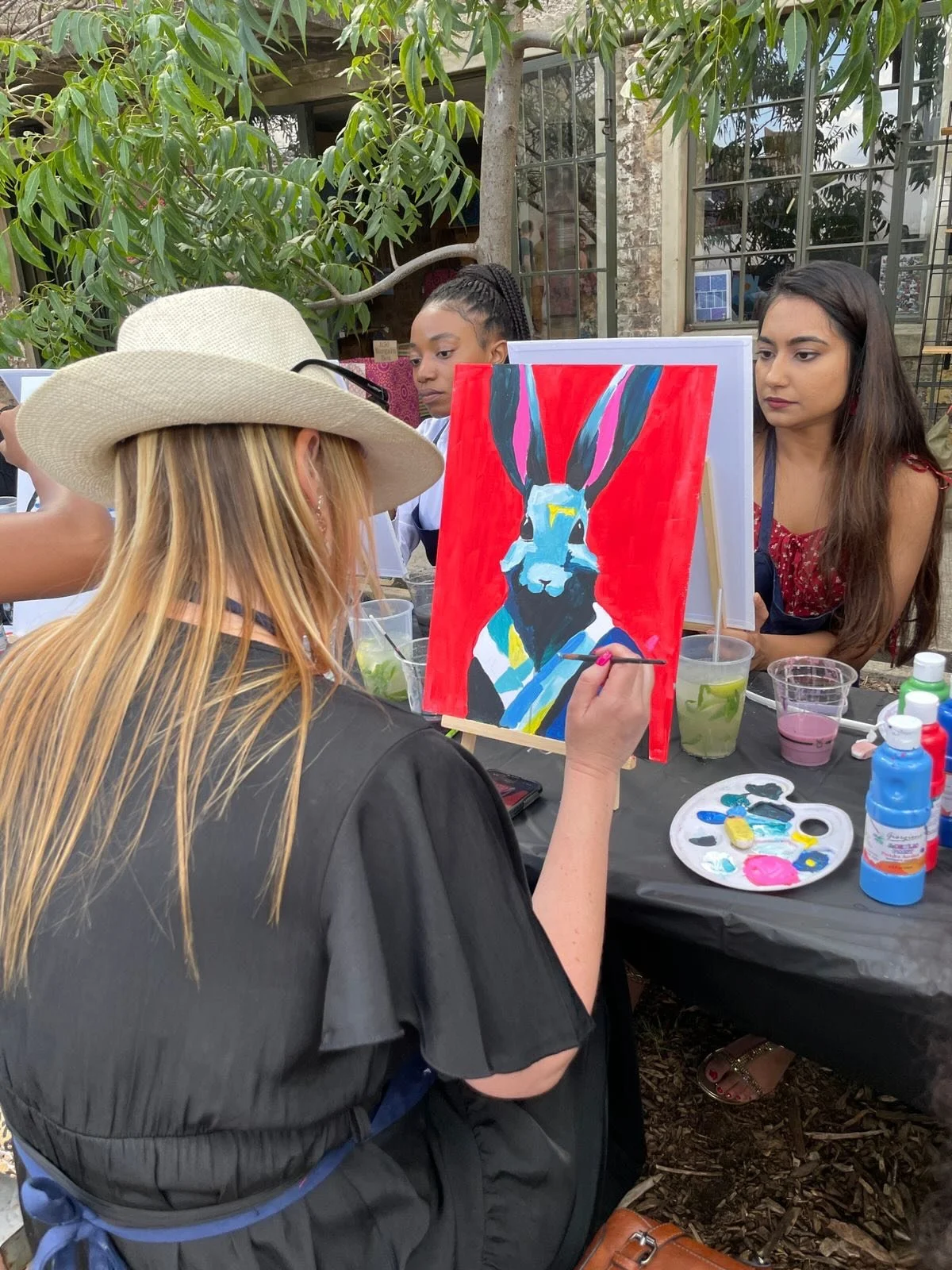 A woman with long blonde hair wearing a wide-brimmed hat and black shirt is painting a colorful portrait of a donkey on a canvas. The portrait features a donkey with bright blue and pink tones on a red background. There are other women observing and 