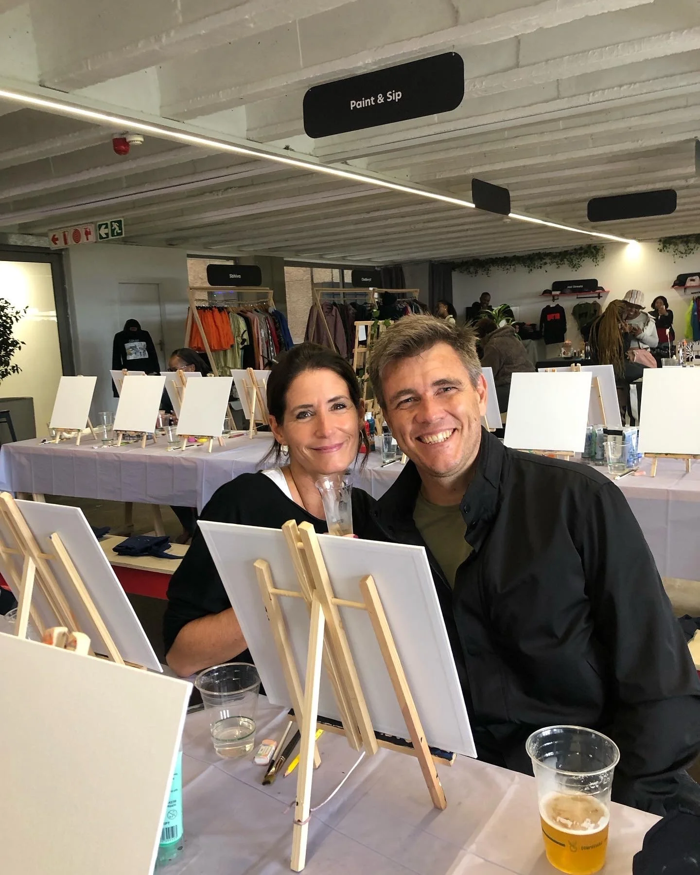 A man and a woman smiling and sitting together at an art event with blank canvases on easels in front of them. In the background, there are clothes on racks and several people, some standing and some sitting, in a well-lit indoor space.