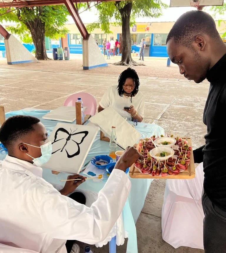 A man offers a tray of appetizers to a person painting a butterfly at an outdoor event under a shelter. A woman is sitting in the background, looking at her phone, with trees and other people in the distance.