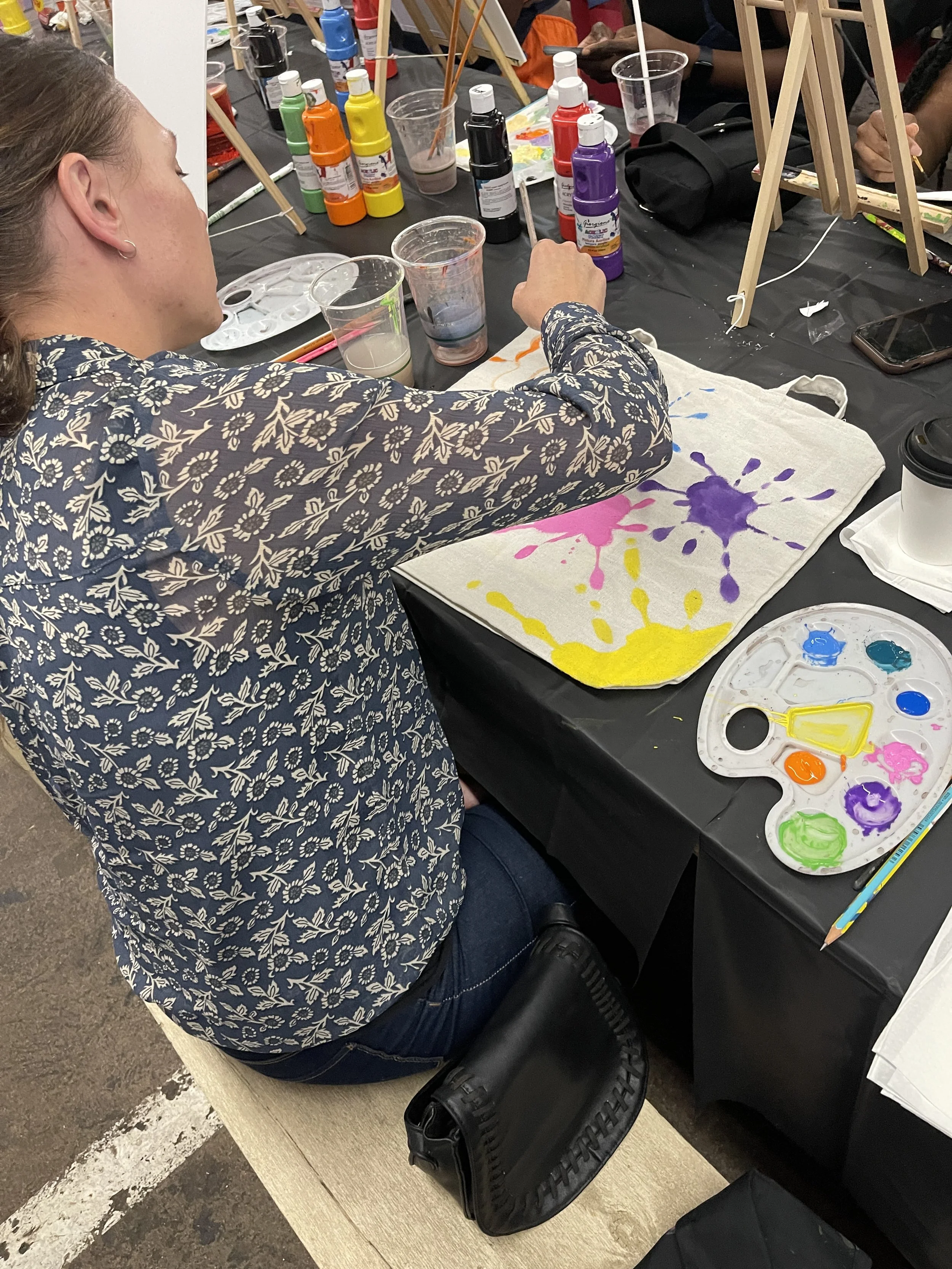 A person sitting at a table with art supplies, including paint bottles, brushes, and a paint palette, engaged in painting on a piece of fabric with colorful splashes. The table also has cups, a phone, and a black bag. The environment appears to be a 