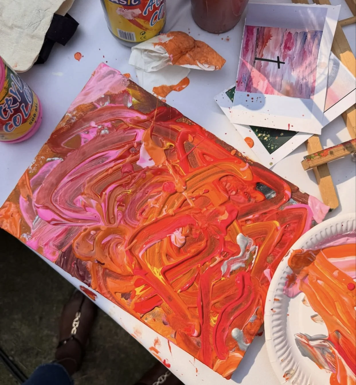 Artwork in progress on a canvas, with bright pink, orange, yellow, and red paint mixed and swirled. Art supplies and paper with painted impressions are visible on the table.