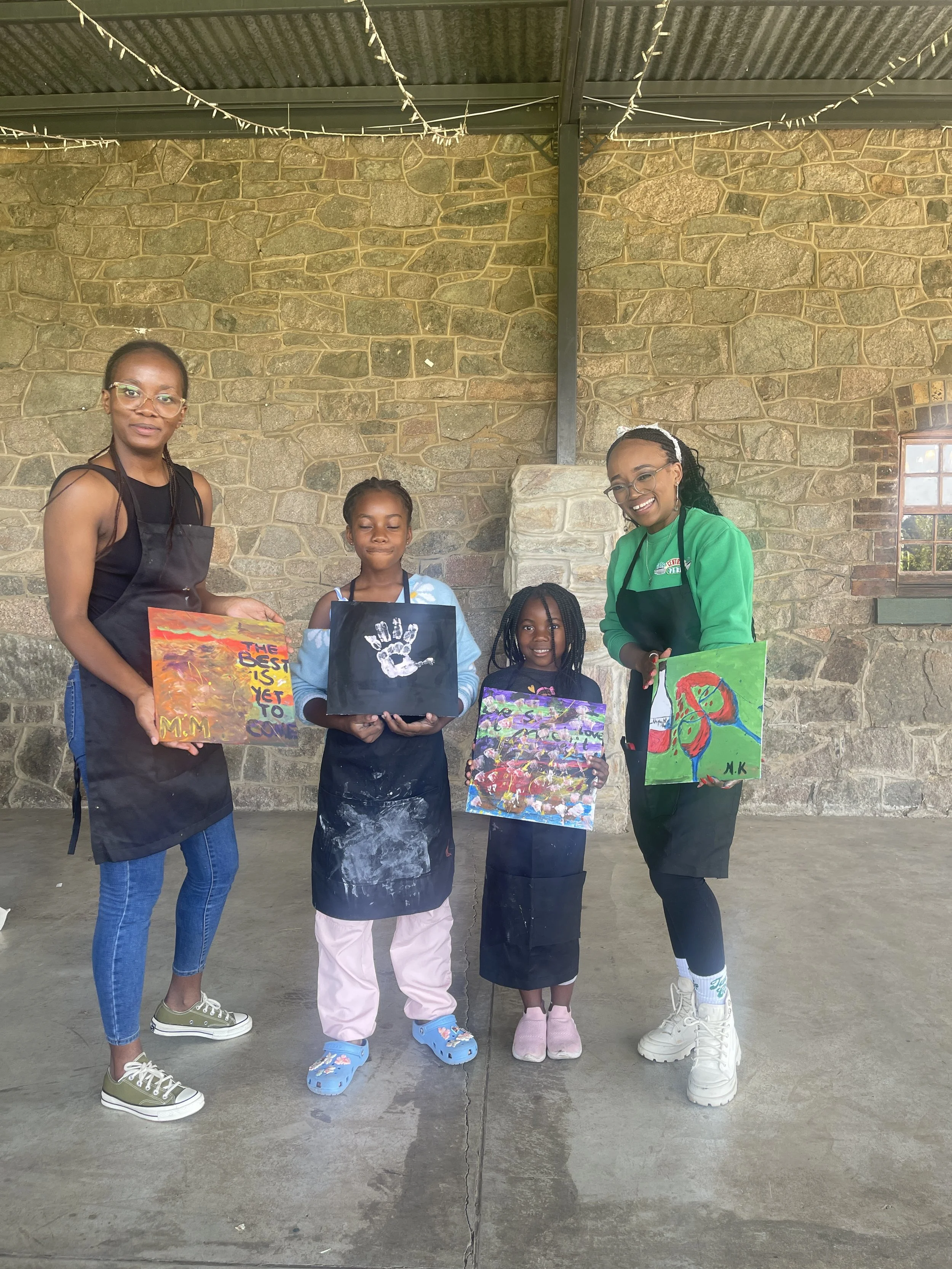 A group of five people standing in front of a stone wall, holding colorful paintings. The group includes three children and two adults, with the children in the middle and the adults on either side. The adults are wearing black aprons and casual clot