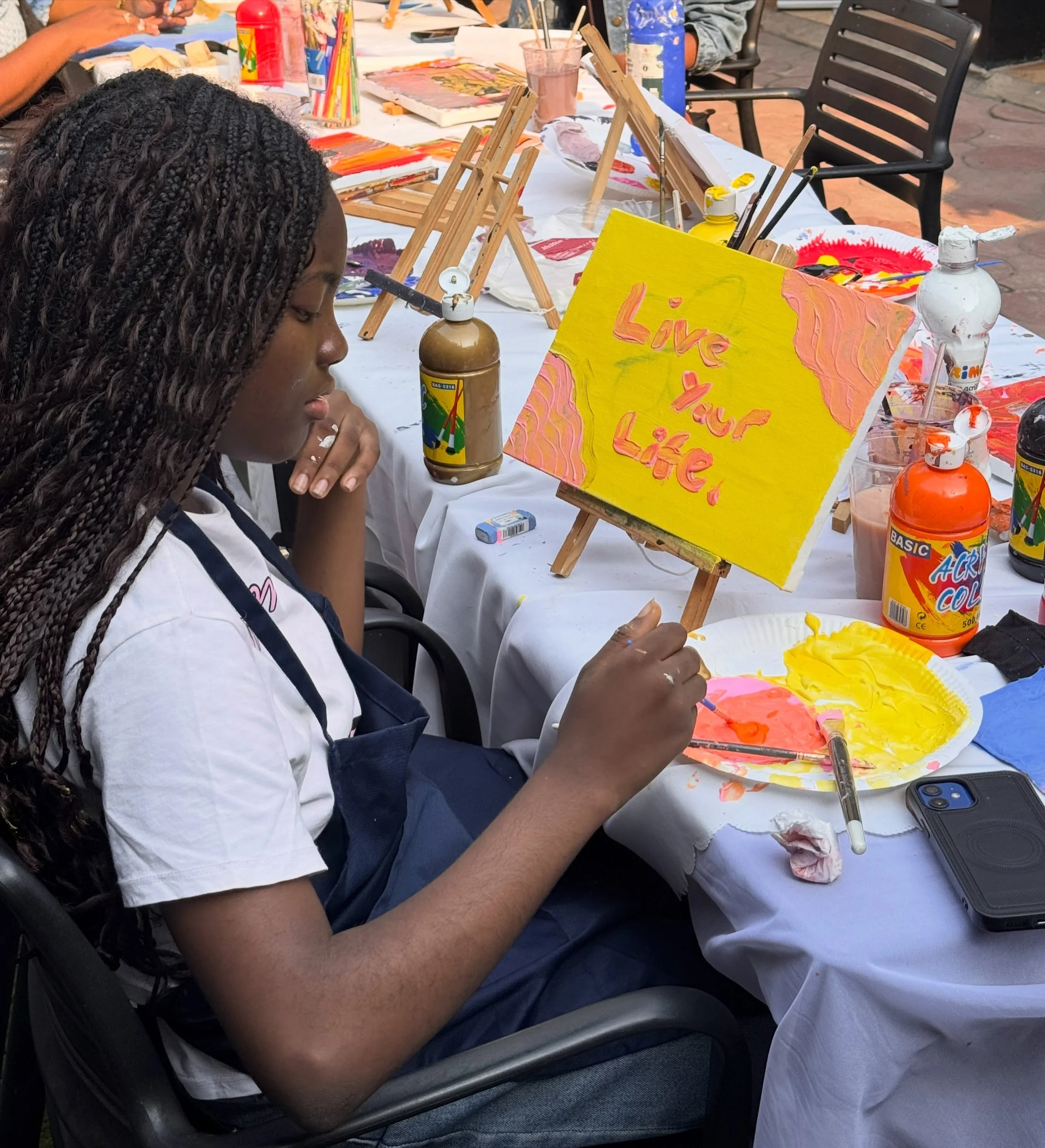 A person with long, braided hair sitting at a table painting a bright yellow canvas with the words 'Live Your Life' in pink. The table is covered with various paints, brushes, and art supplies, and other people are visible in the background also pain