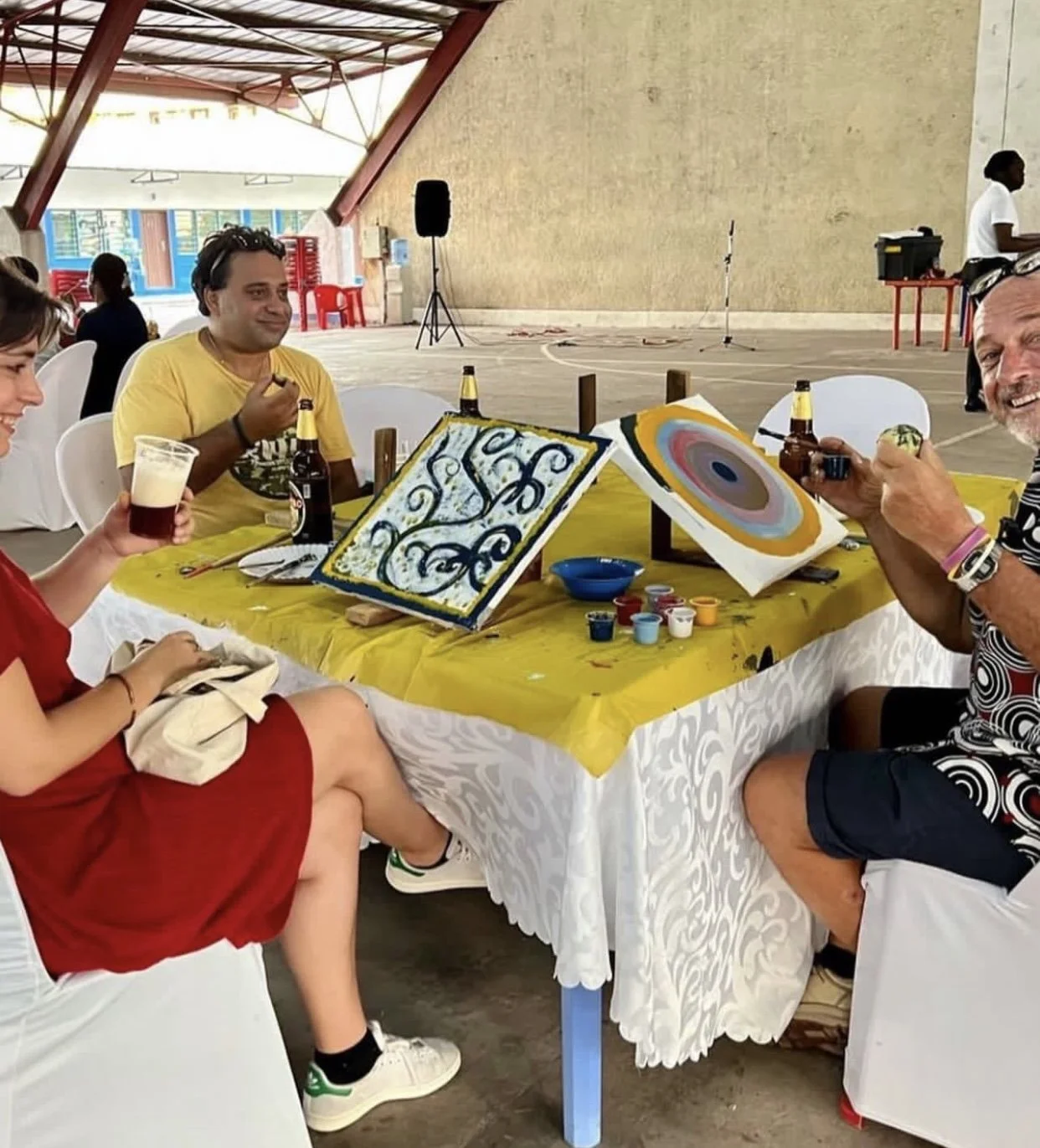 Group of people sitting around a table with painting supplies, chocolate beer bottles, and painted canvases, enjoying drinks and art in an indoor setting.