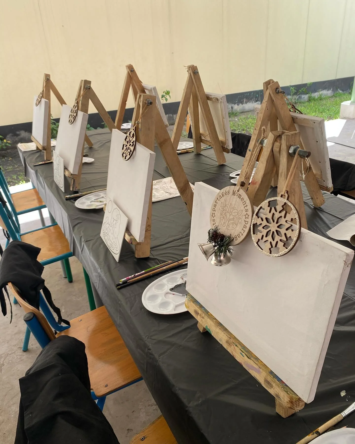 Table with painting supplies, including small blank canvases on wooden easels, paint palettes, brushes, and holiday-themed wooden ornaments, set up for a painting event outdoors.