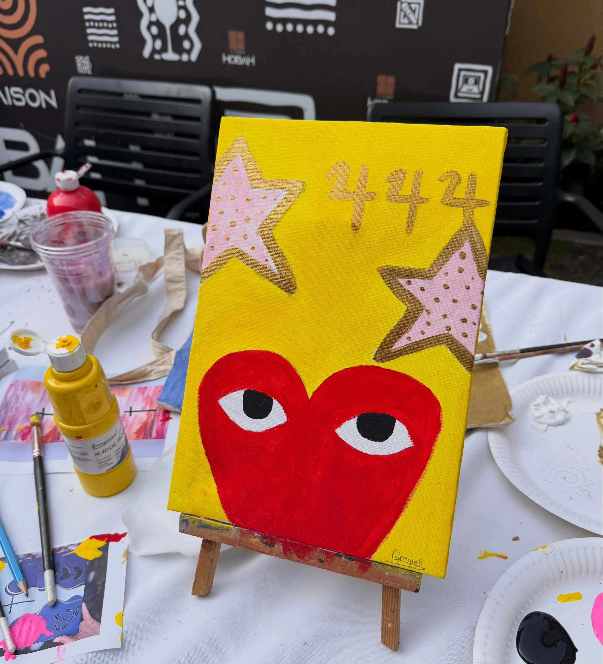 A small craft display with a painting of Elmo the puppet from Sesame Street, featuring a big red face and wide eyes, on a yellow canvas. The background includes art supplies such as paint bottles, brushes, and paper, and a black cardboard display boa