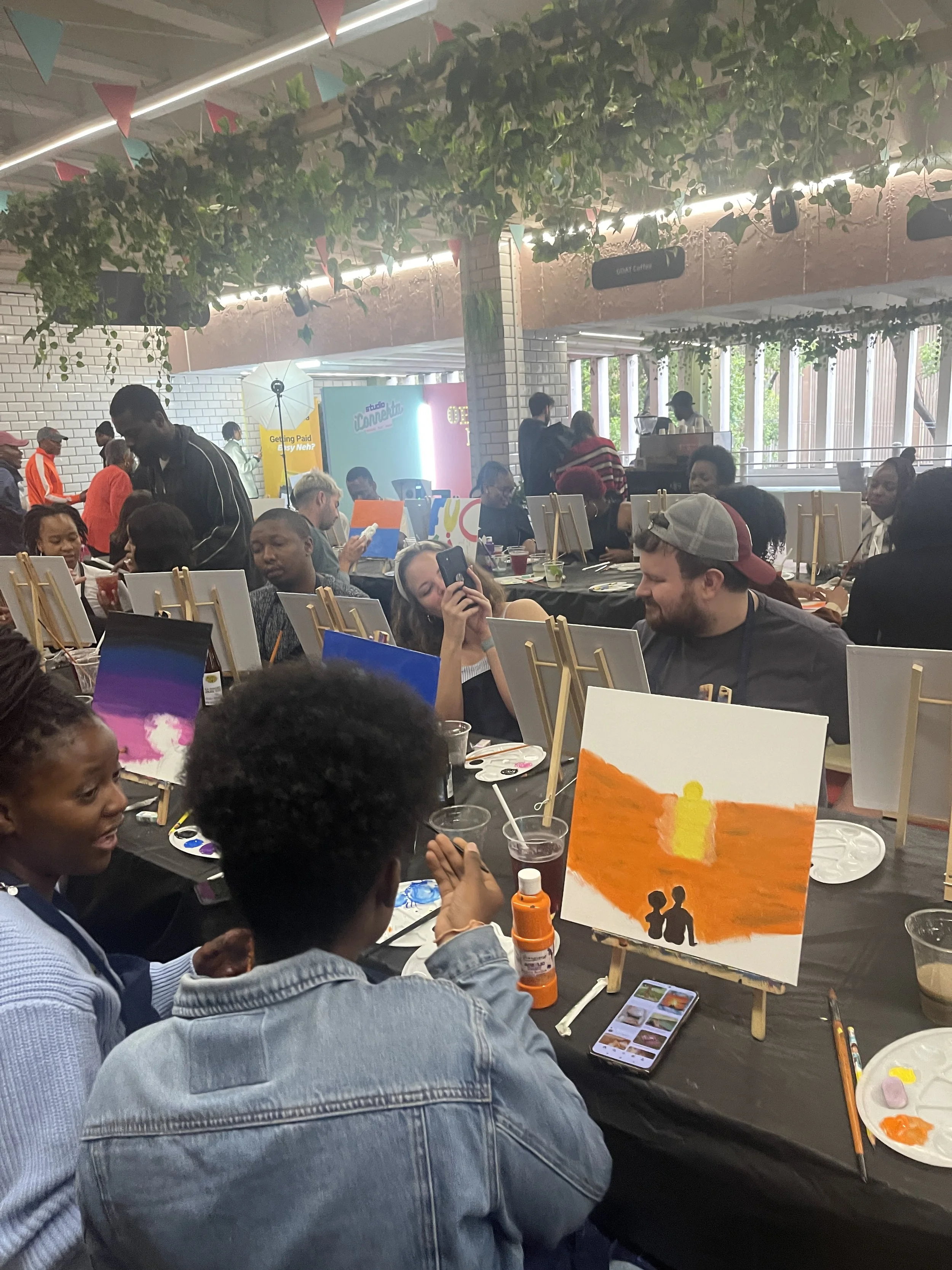 A group of diverse people participating in a painting workshop at a table decorated with art supplies, paintings, and refreshments, with greenery and colorful banners overhead.