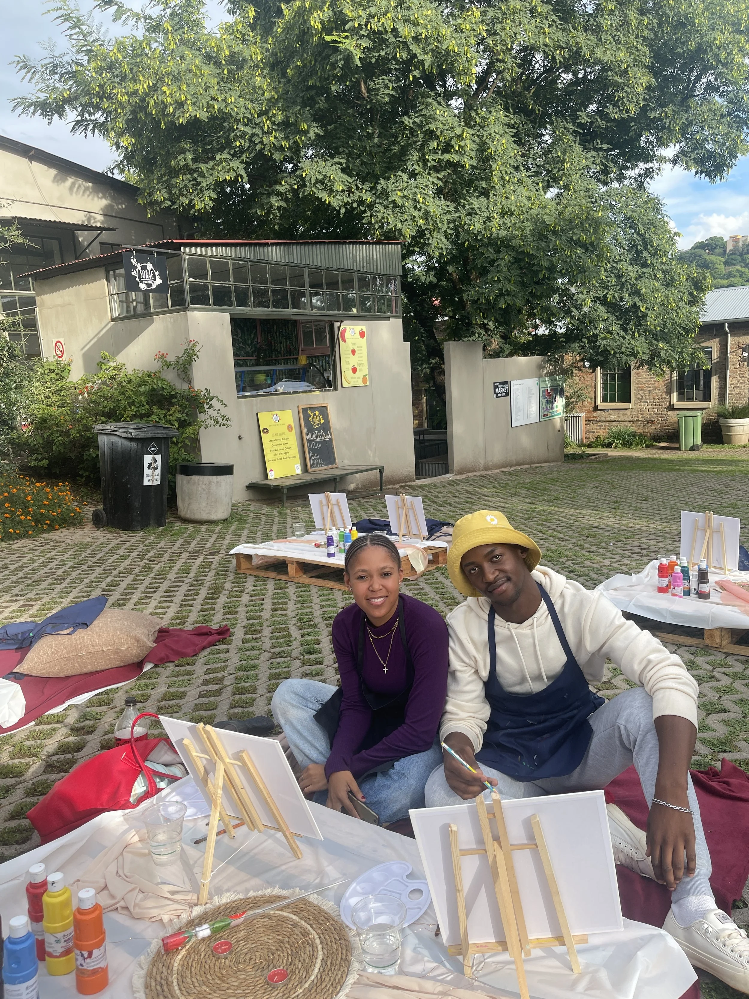 Two people sitting on the ground at a paint and sip event outdoors. There are art supplies, small canvases on easels, and bottles of paint on a table in front of them. A large tree and buildings are in the background.