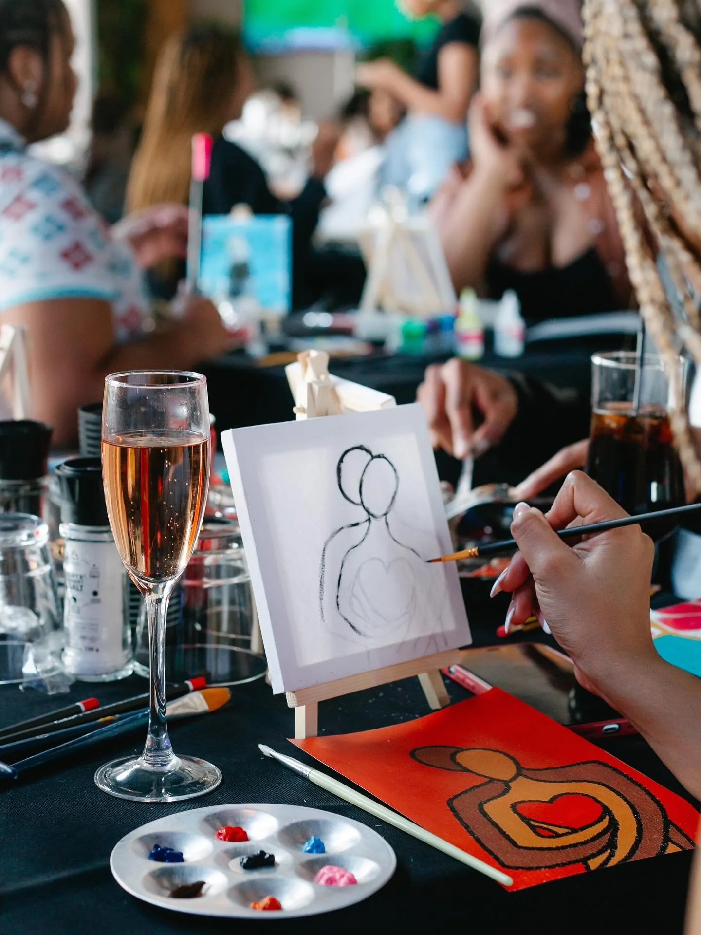 A woman is painting a colorful abstract art piece, featuring shapes in orange, red, yellow, and brown, on a small canvas. She is holding a paintbrush and working at a table with various art supplies, including a palette with paint and a glass of rosé wine. In the background, there are other people engaging in creative activities at a social event.