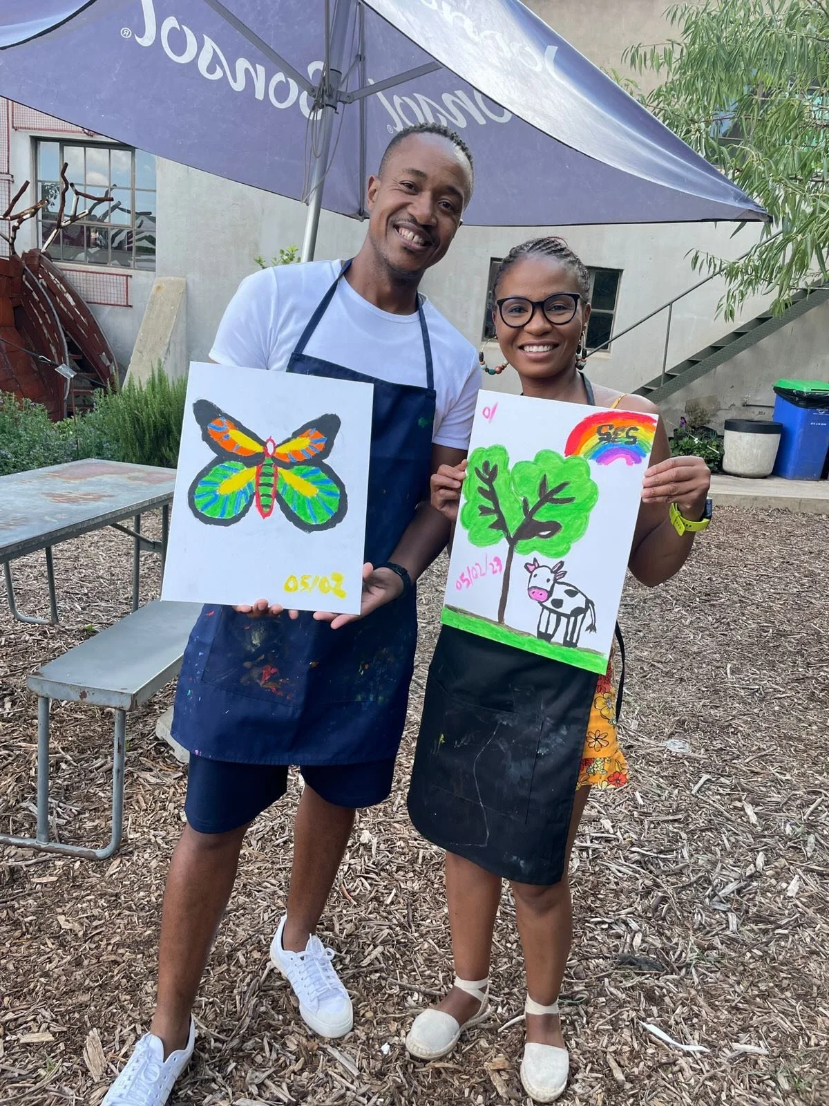A man and woman standing outdoors underneath a large purple umbrella, each holding colorful artwork. The man's drawing is of a brightly colored butterfly, and the woman's drawing features a tree, a cow, and a rainbow. They are smiling and wearing cas