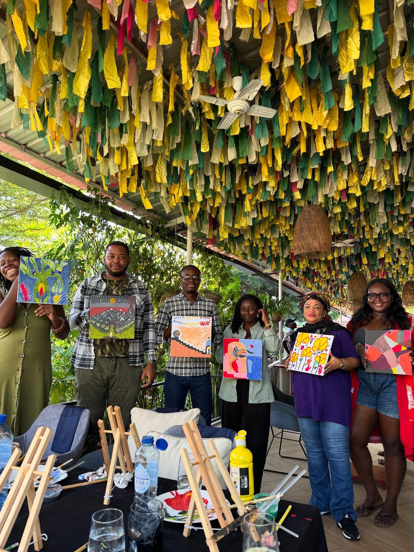 Group of people holding colorful paintings in a decorated outdoor setting, with art supplies and water bottles on a table in the foreground.