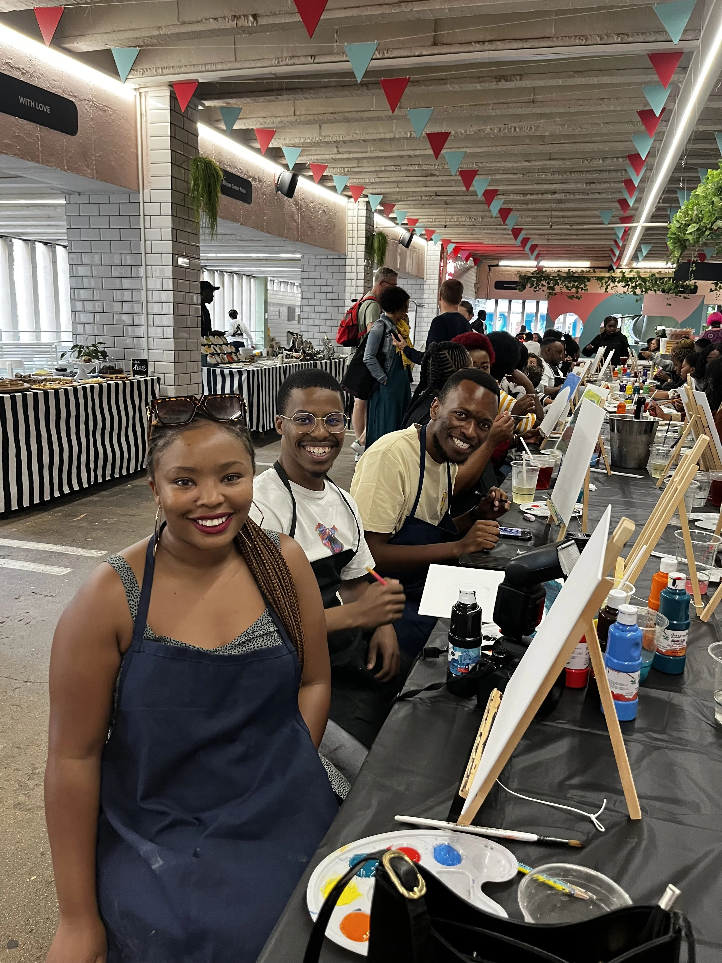 People painting at a table in an indoor art studio with masks of paints, brushes, and canvases. Several people are smiling and engaging in painting activities.
