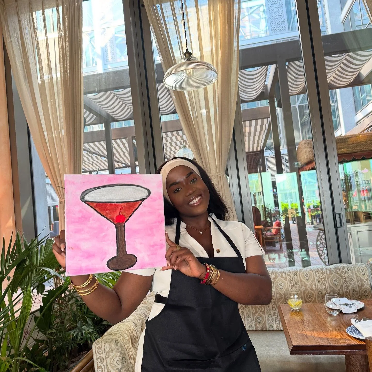 A woman in a restaurant holding a painting of a cocktail glass with a red cherry, smiling at the camera.