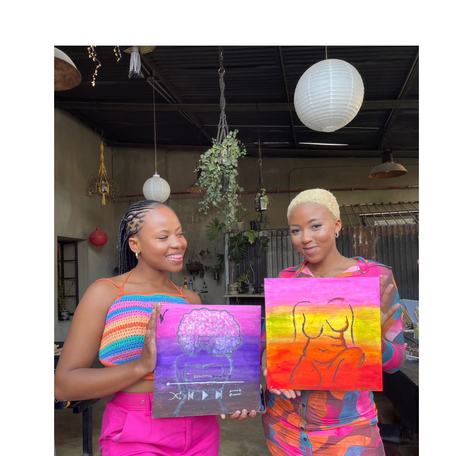 Two women smiling and holding colorful paintings of women with curly hair, in a room with hanging paper lanterns, plants, and rustic decor.