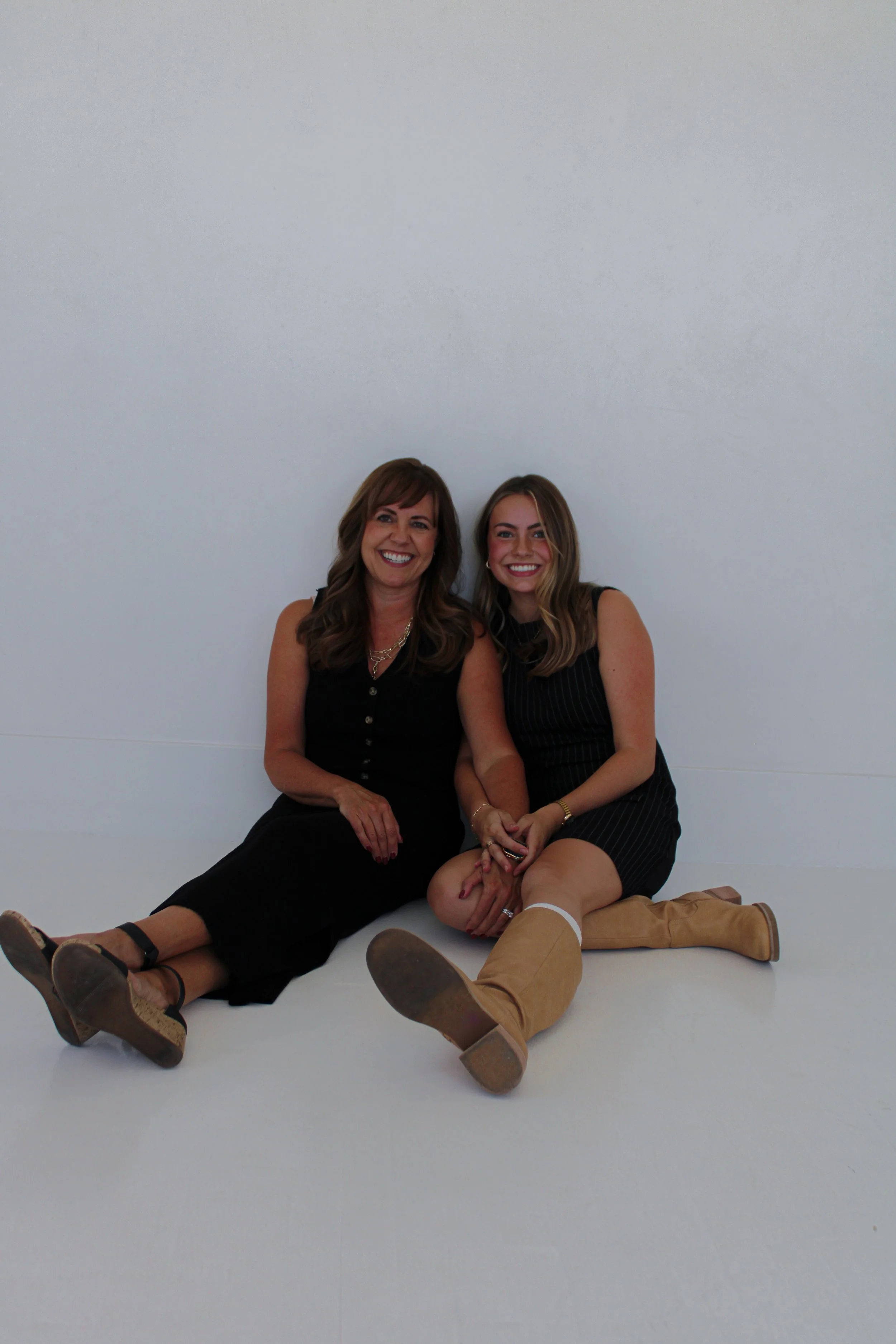 Two women sitting on the floor against a white wall, smiling, with their legs crossed and holding hands, dressed in black outfits and brown boots.