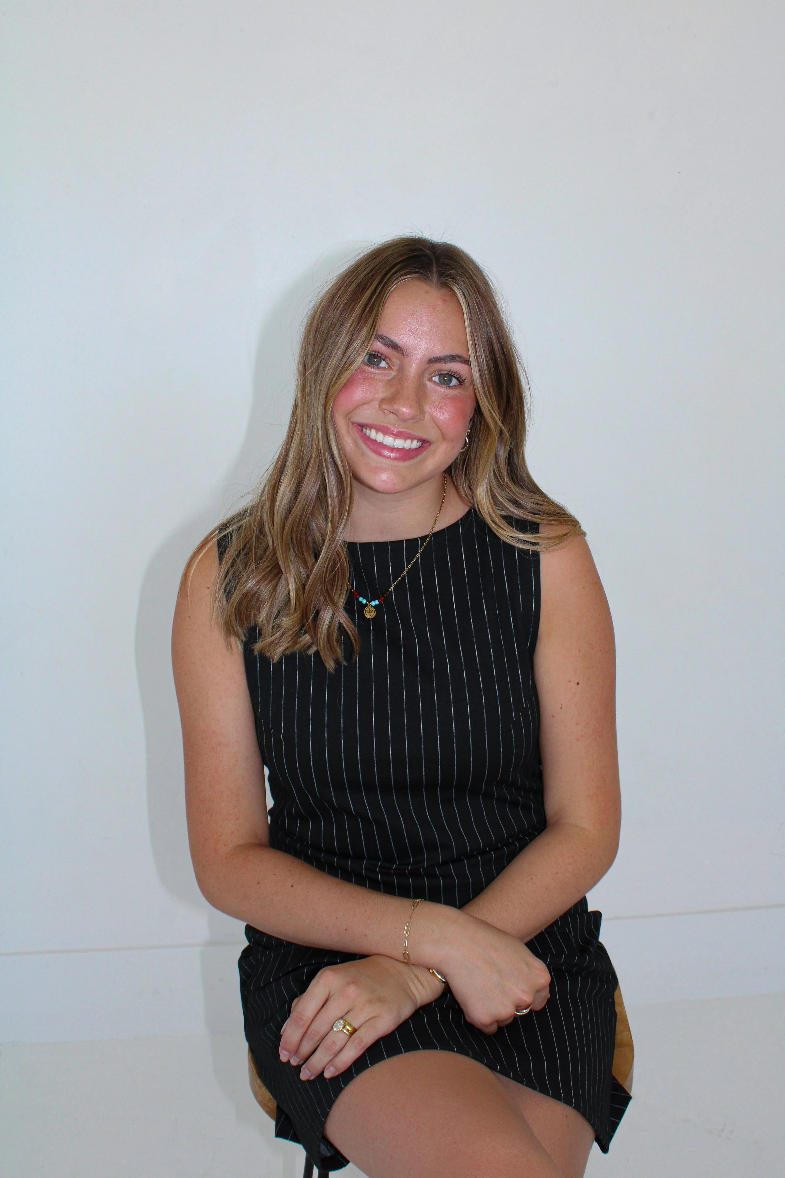 A young woman with long, wavy brown hair, smiling, seated against a plain white wall. She is wearing a sleeveless black dress with white pinstripes, gold jewelry including rings, a necklace, and a bracelet.