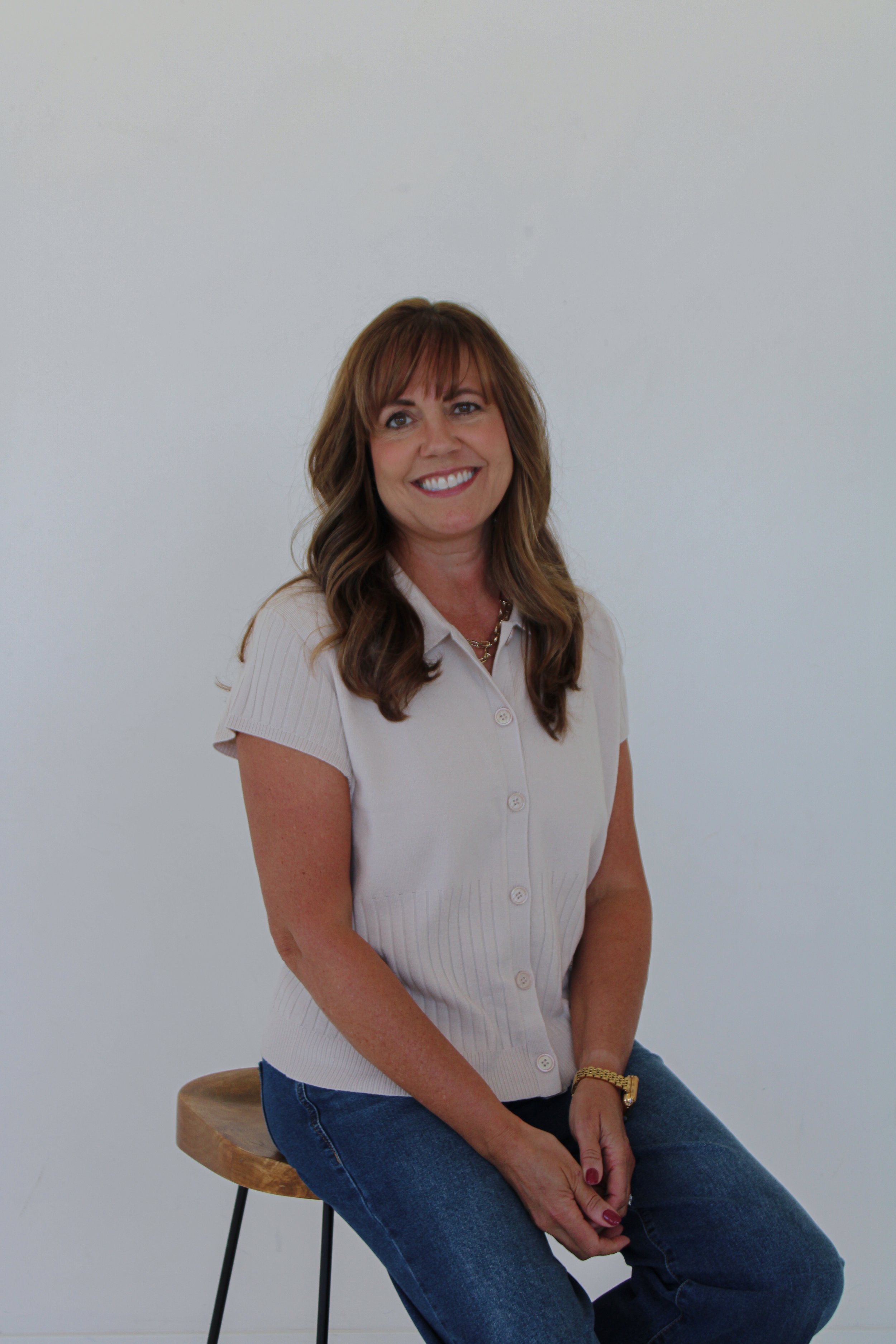 A smiling woman with brown hair, wearing a beige short-sleeved button-up shirt and blue jeans, sitting on a wooden stool with a black metal leg against a plain white wall.