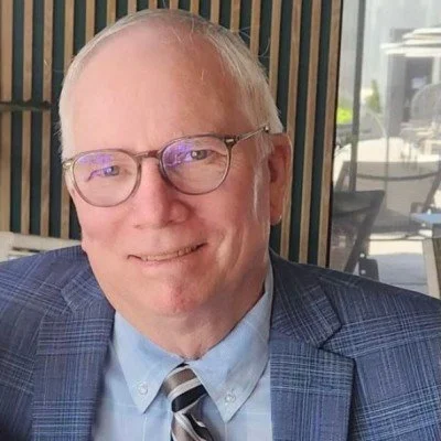 A smiling elderly man with glasses, wearing a blue plaid blazer, light blue shirt, and a striped tie, sitting in a bright indoor setting with a window and outdoor scenery in the background.