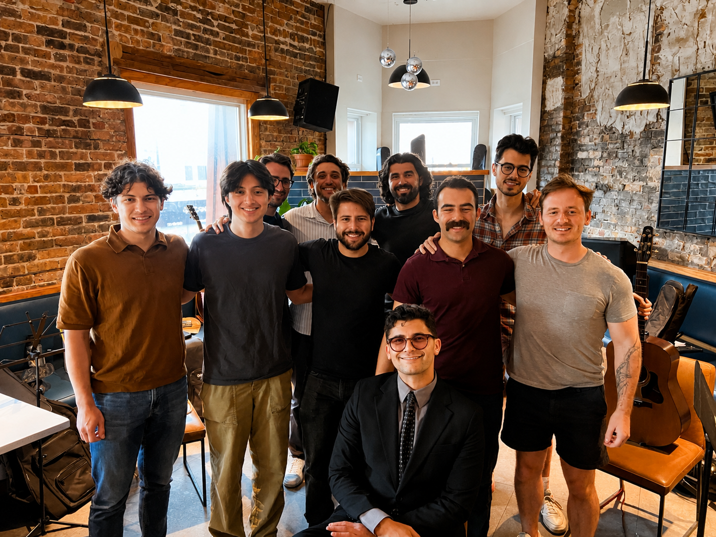 A group of ten smiling young men in casual and formal clothing posing together in a room with brick and concrete walls, musical instruments, and equipment.