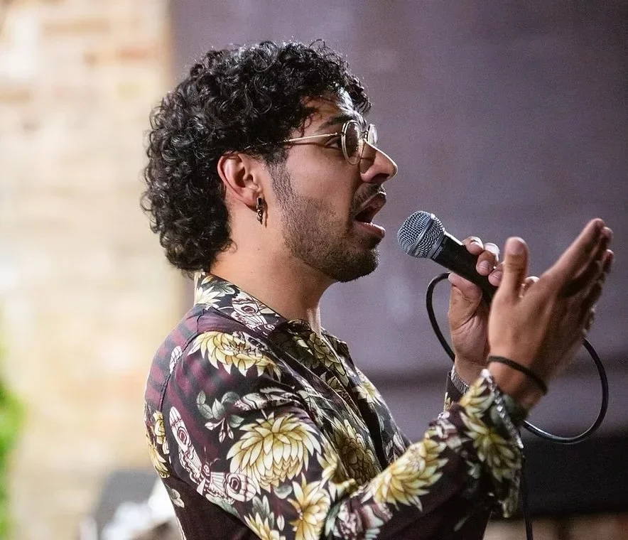 A man with curly hair, glasses, earrings, and a floral shirt speaking or singing into a microphone. Voice & Trumpet Teacher Ricardo Jimenez.