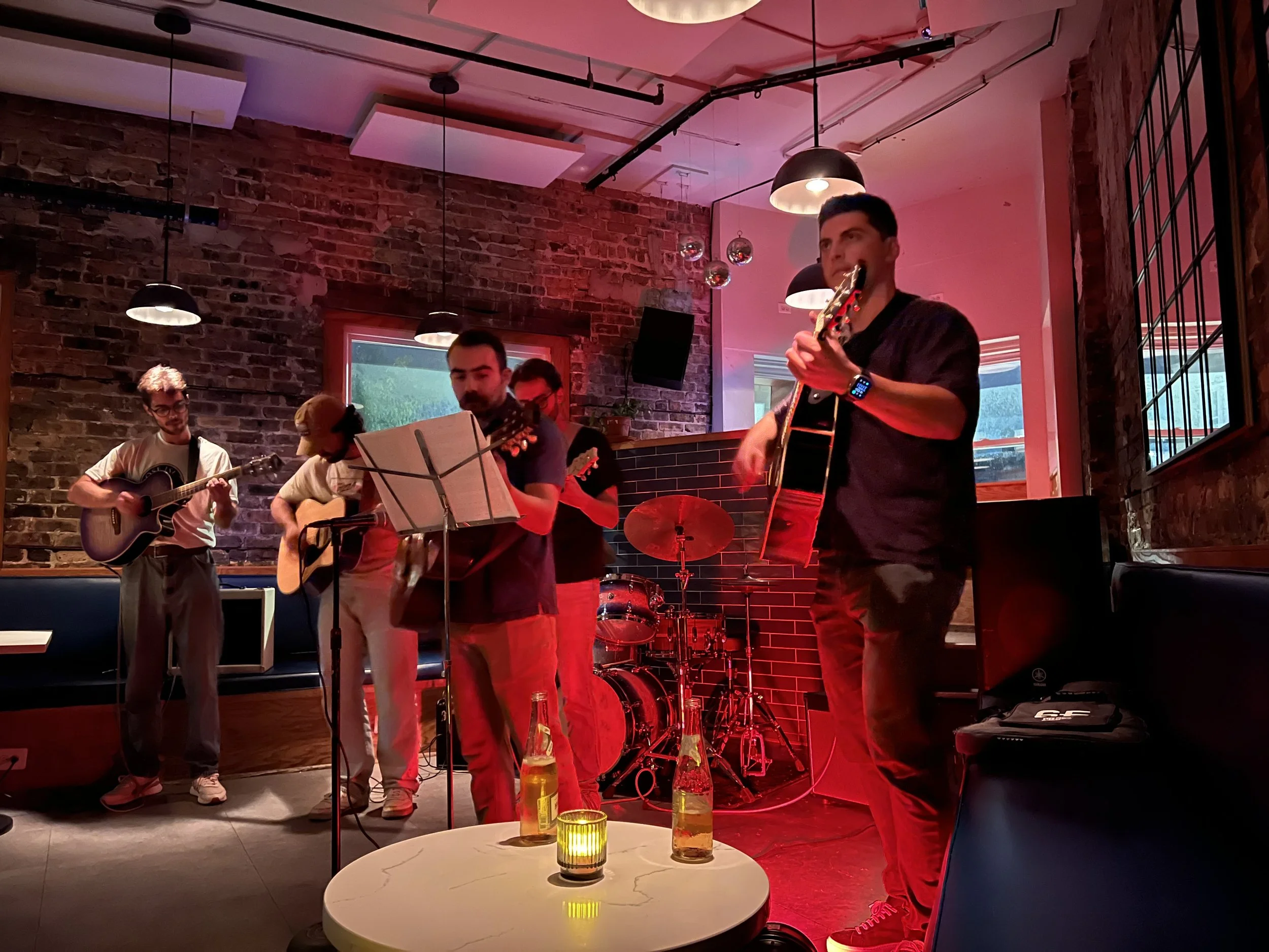 A group of musicians performing in a cozy, dimly lit venue with brick walls and hanging lights. There are four men with guitars, and a drum set in the background. Two beers and a candle are visible on a small round table in the foreground.
