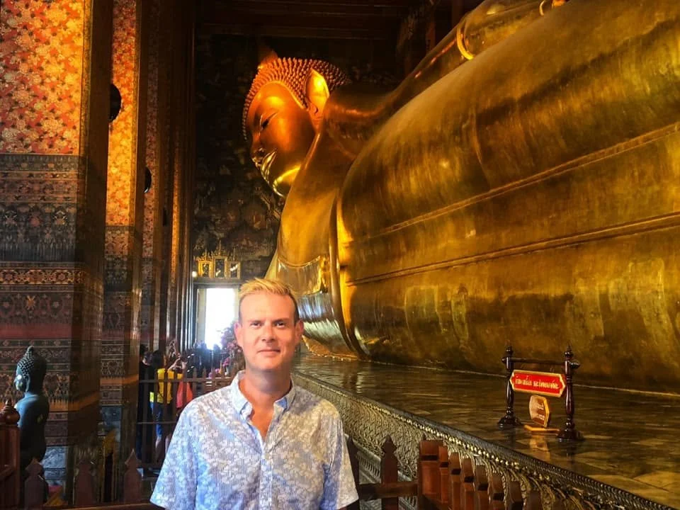 A man standing in front of a large reclining golden Buddha statue inside a temple with intricate wall art.