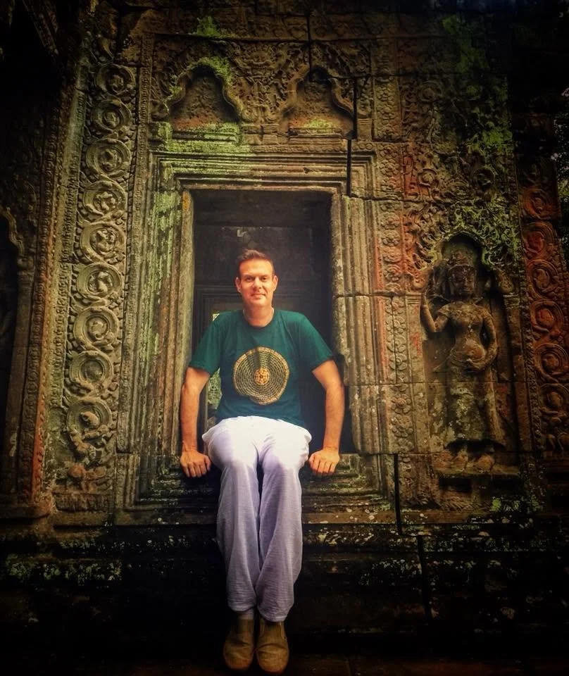 A young man sitting on the stone window ledge of an ancient, carved stone temple or structure, with intricate designs and relief sculptures, including a figure of a woman on the right side.