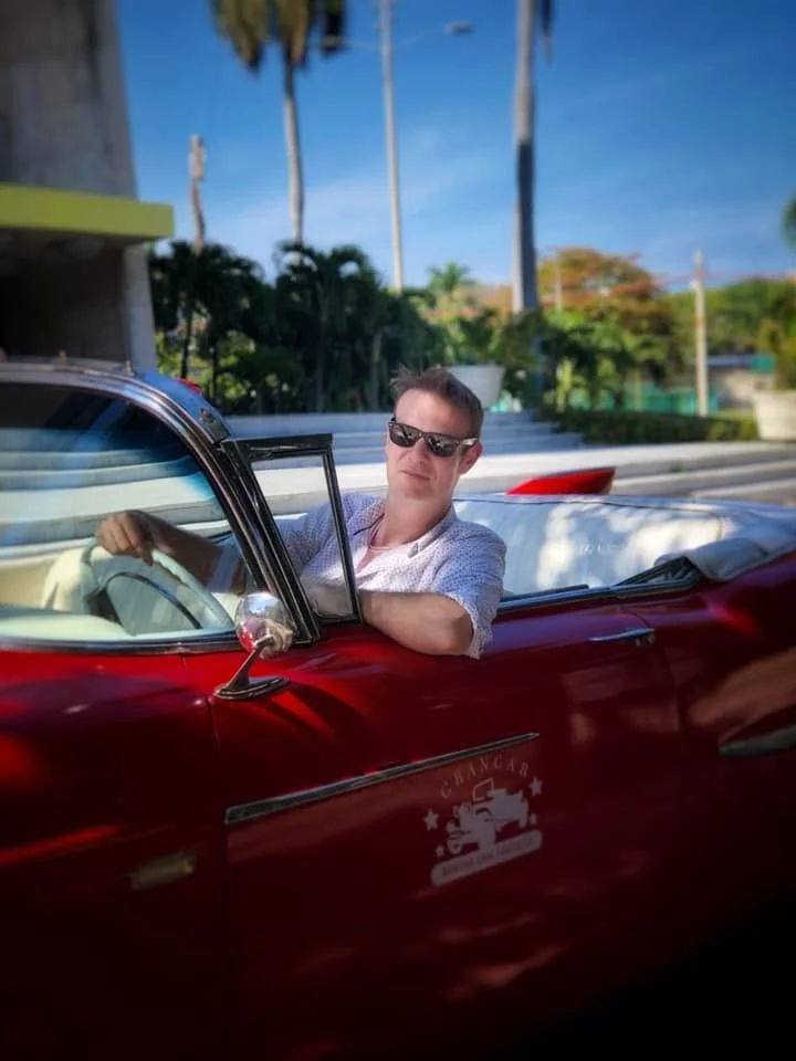 A man wearing sunglasses sitting in a vintage red convertible car with a white interior on a sunny day with palm trees in the background.