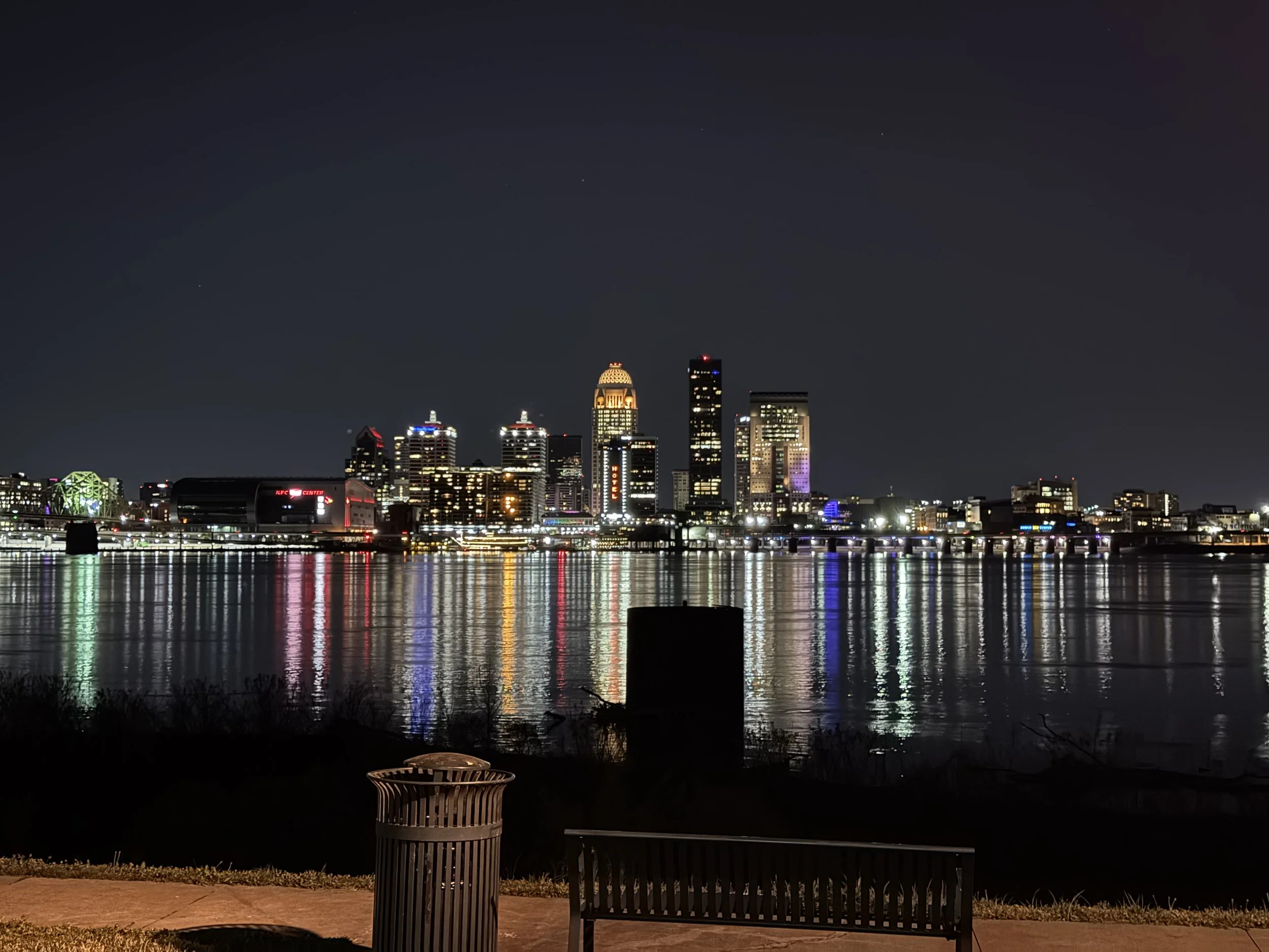 Nighttime city skyline with illuminated buildings reflected on the water, viewed from a park area with benches and a trash can in the foreground.