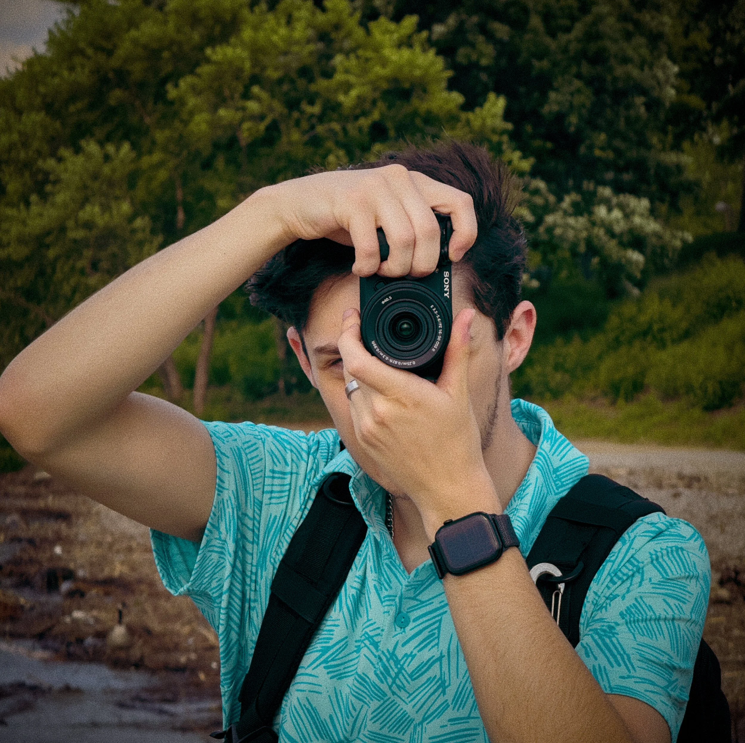 Person taking a photo with a Sony camera outdoors surrounded by green trees, wearing a blue patterned shirt, a black watch, and a ring on their finger.